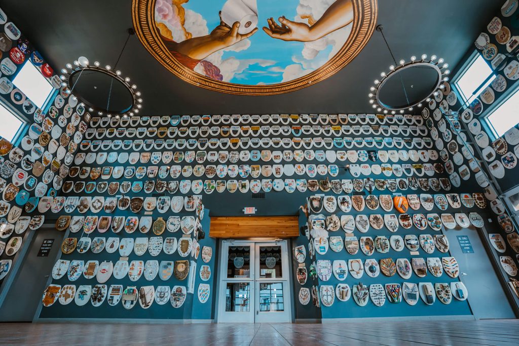 An endless array of decorated toilet seats cover an entire wall floor to ceiling. Two circular chandeliers hang on opposite sides of a ceiling fresco that depicts Michelangelo's Creation of Adam hands, where they are offering a roll of toilet paper.