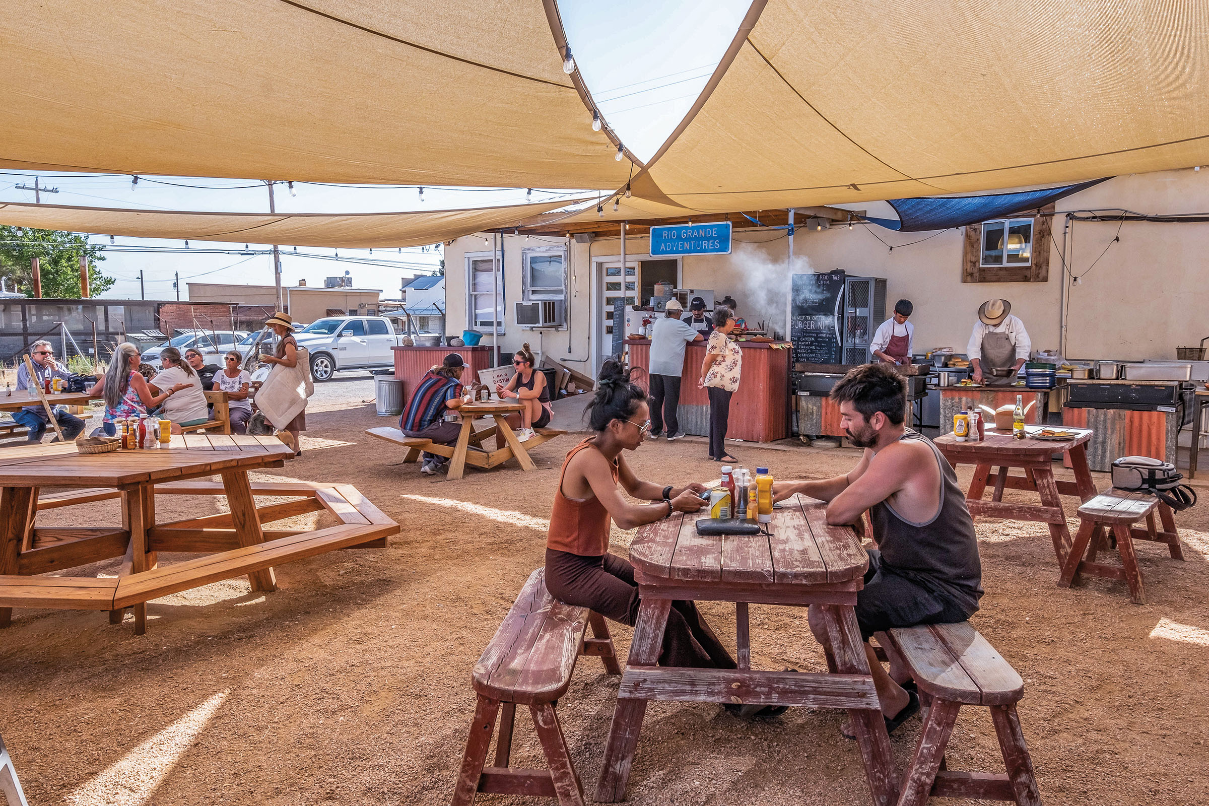 People sit outside at picnic tables under a tan sun shade