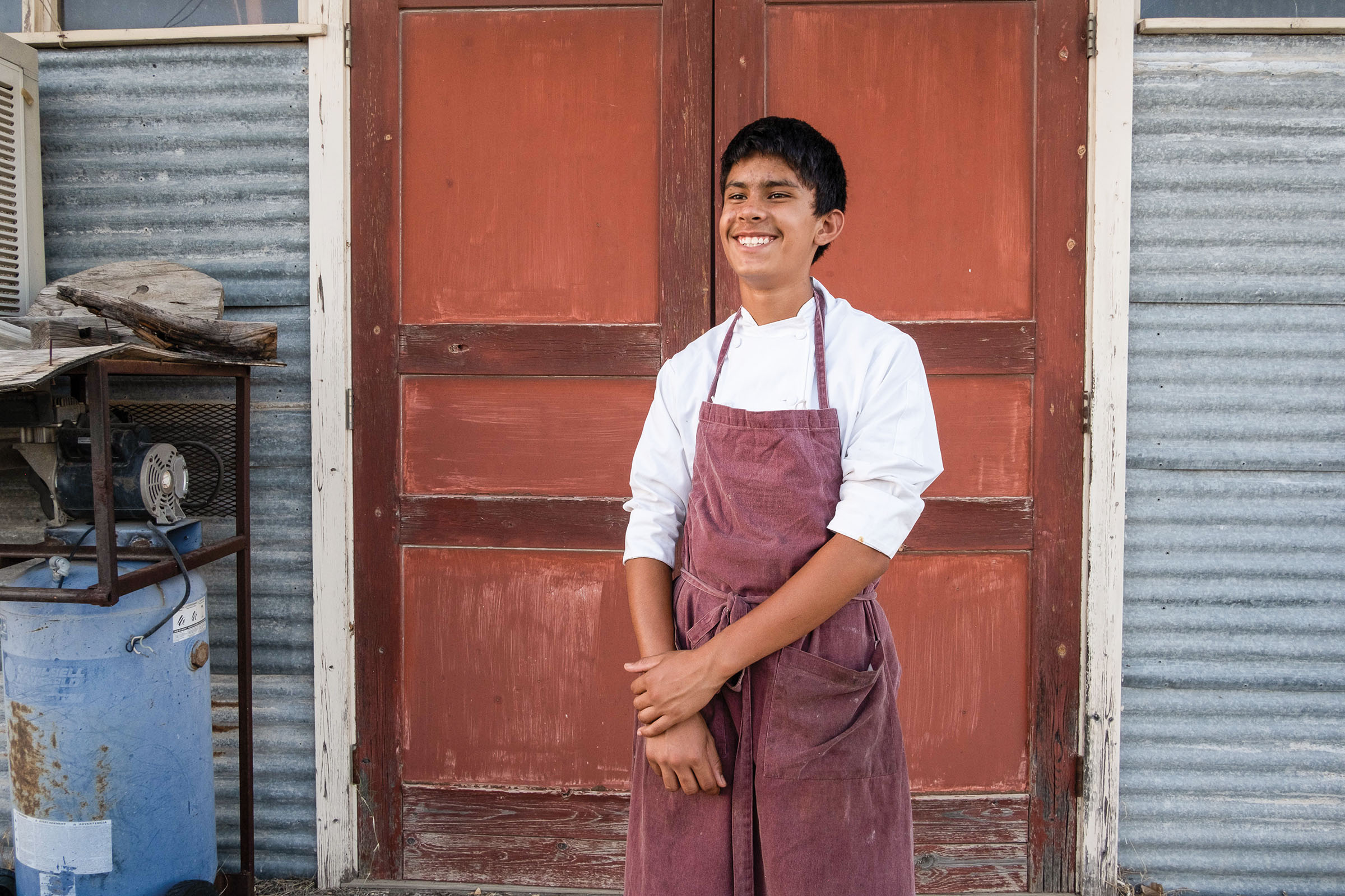 A person in a crisp white shirt and maroon apron smiles with their hands folded in front of a large red door