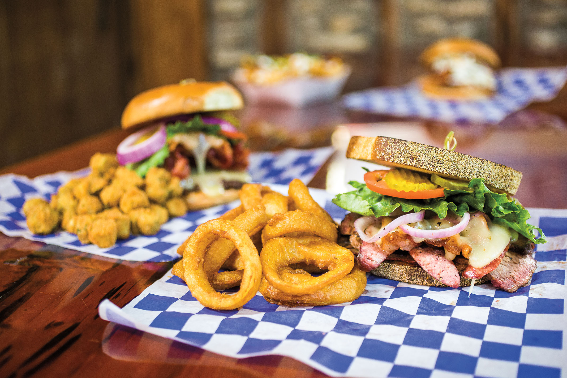 Sandwiches piled high with meat and cheese alongside fried sides like onion rings served on blue checkered paper