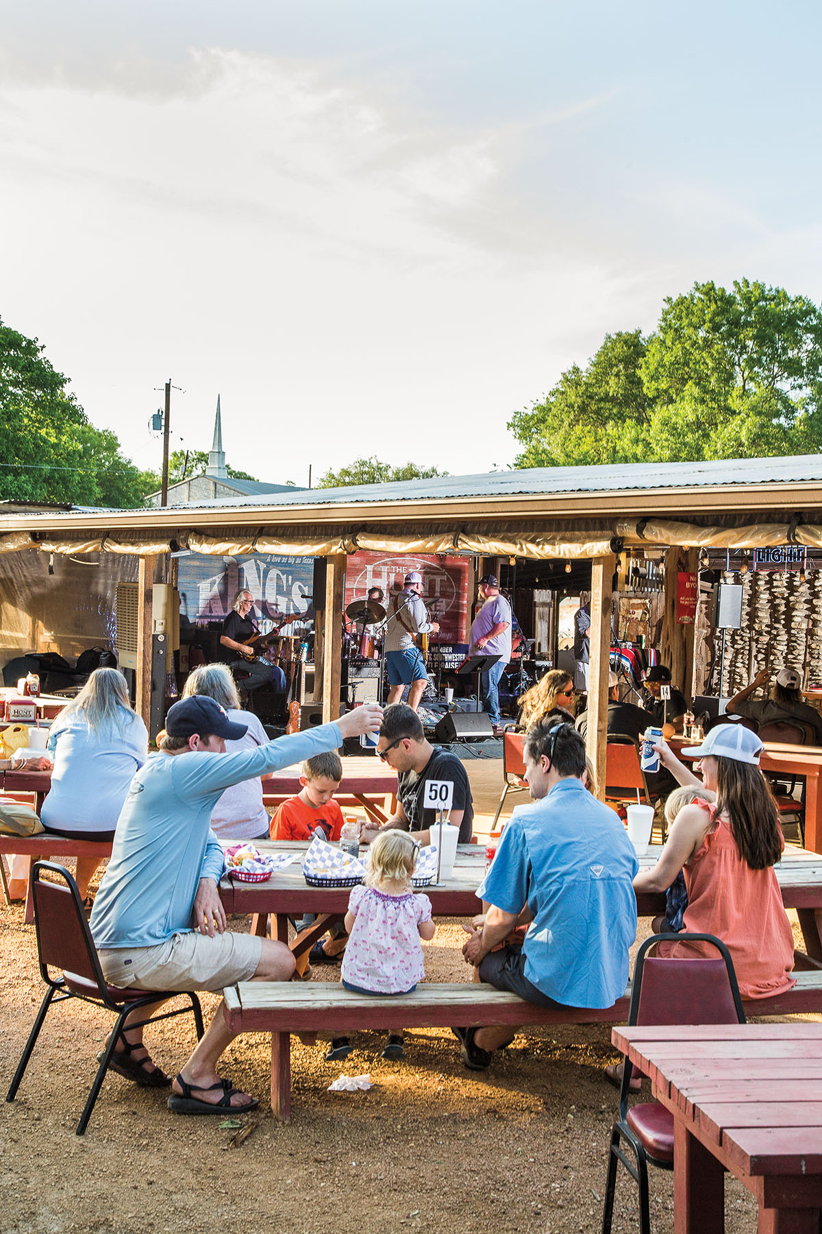 A large group of people sit at outdoor tables near a stage covered by a wooden awning