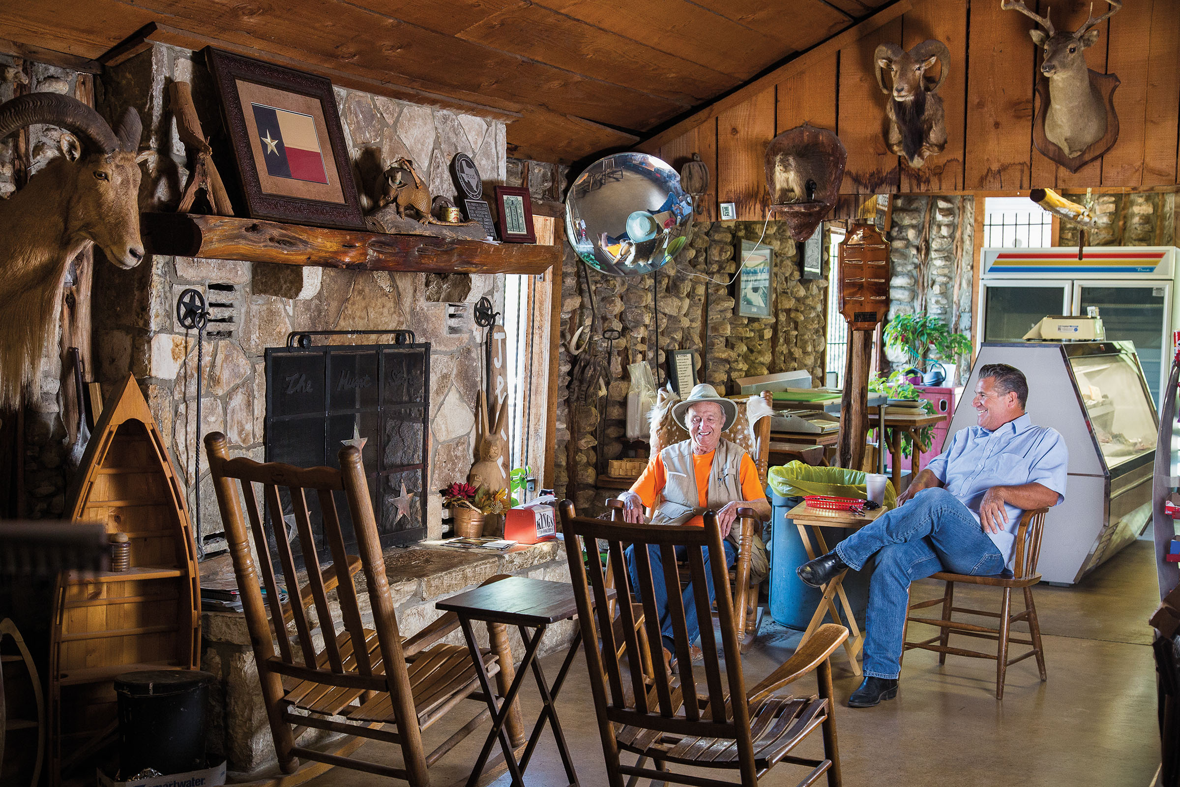 Two men sit in rocking chairs next to a large wooden fireplace in a richly decorated room featuring taxidermy and other items