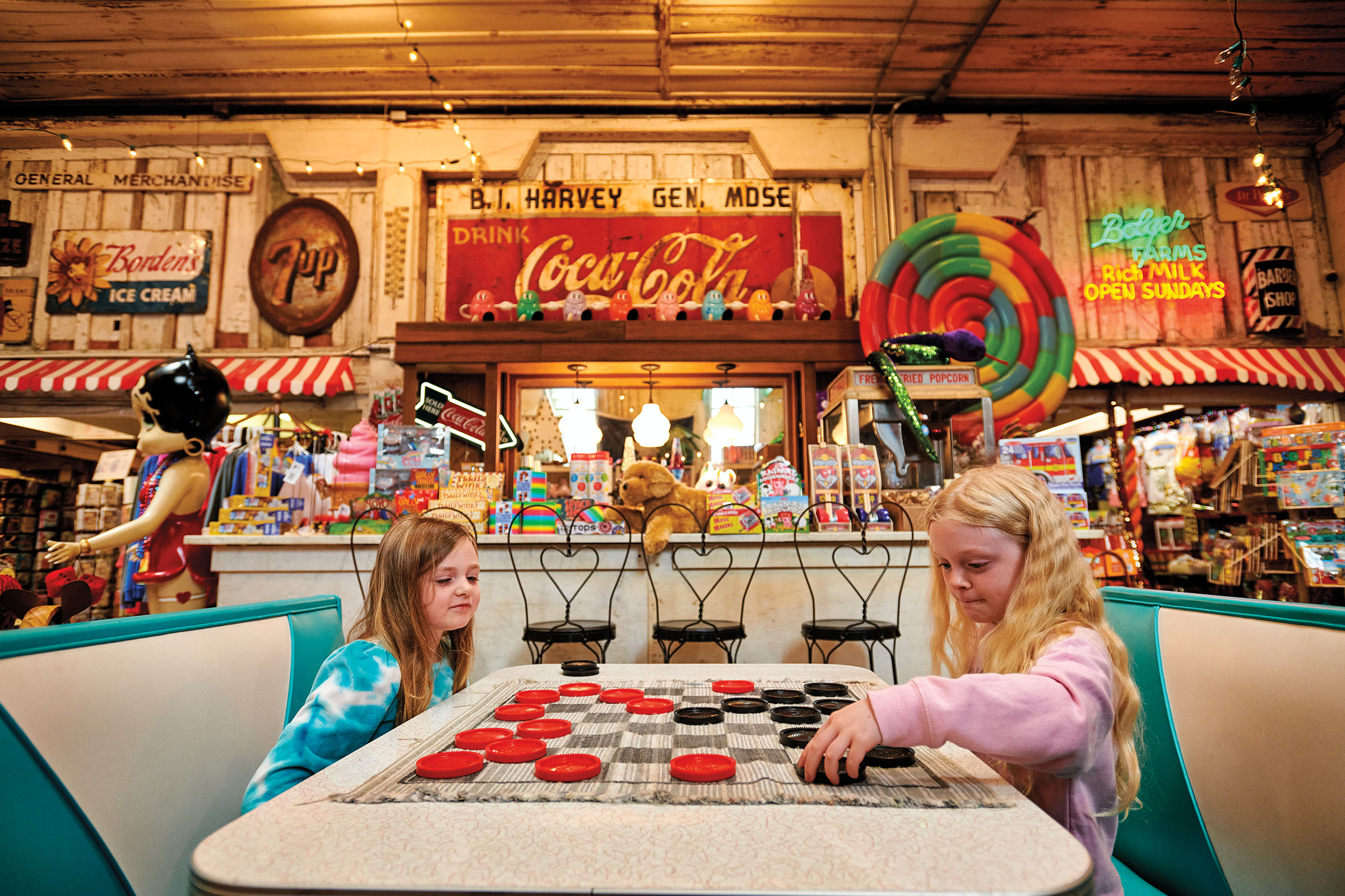 Two young women play checkers at a table in front of a brightly decorated store featuring a vintage Coca-Cola sign