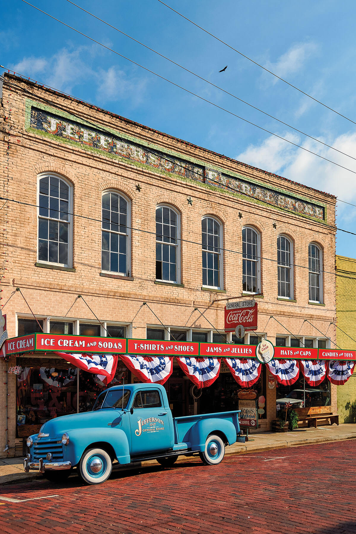 A bright blue vintage pickup truck parked out front of a brick building with red white and blue bunting hanging from the rafters