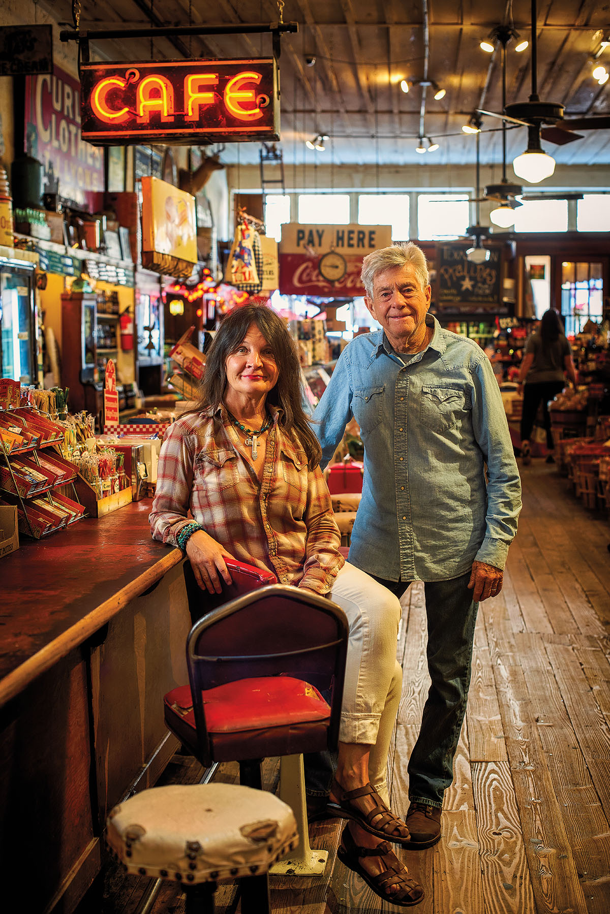 A woman sits at a barstool and a man stands next to her inside of a busy cafe with a red neon sign reading "Cafe"