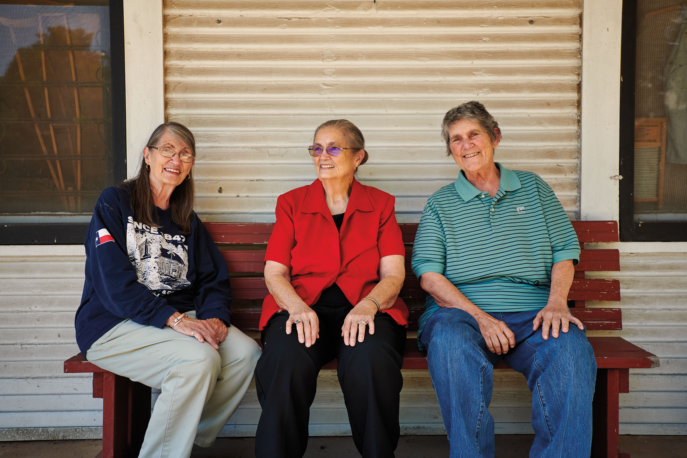 Three women of three generations sit on a wooden bench outside of a building