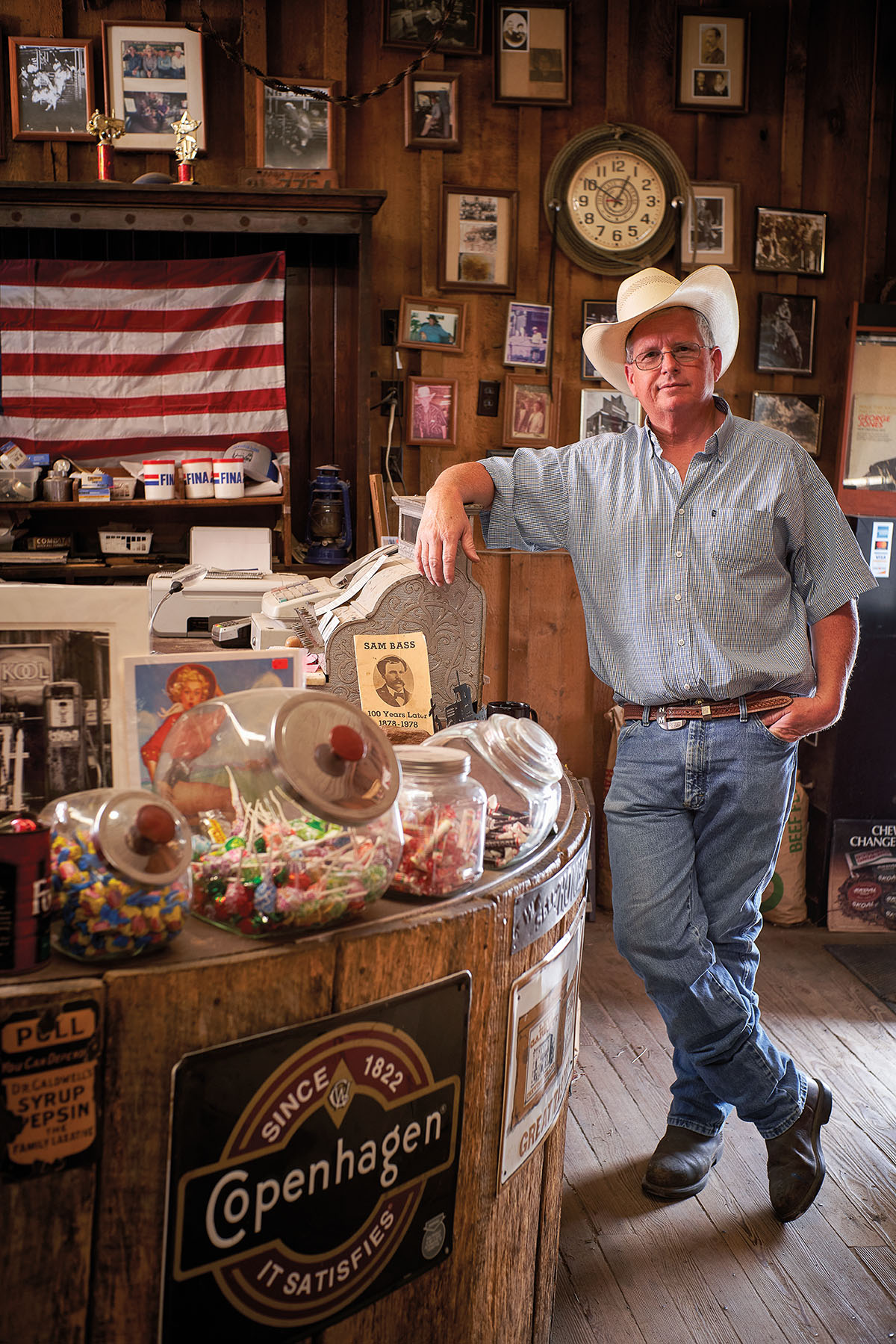 A man in a cowboy hat leans against a busy countertop in front of an American flag