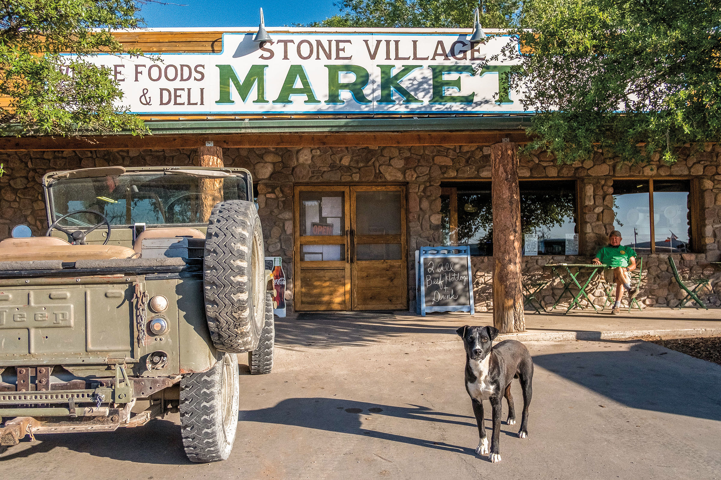 A dog stands next to a vintage jeep outside of a store with a sign reading "Stone VIllage Market"