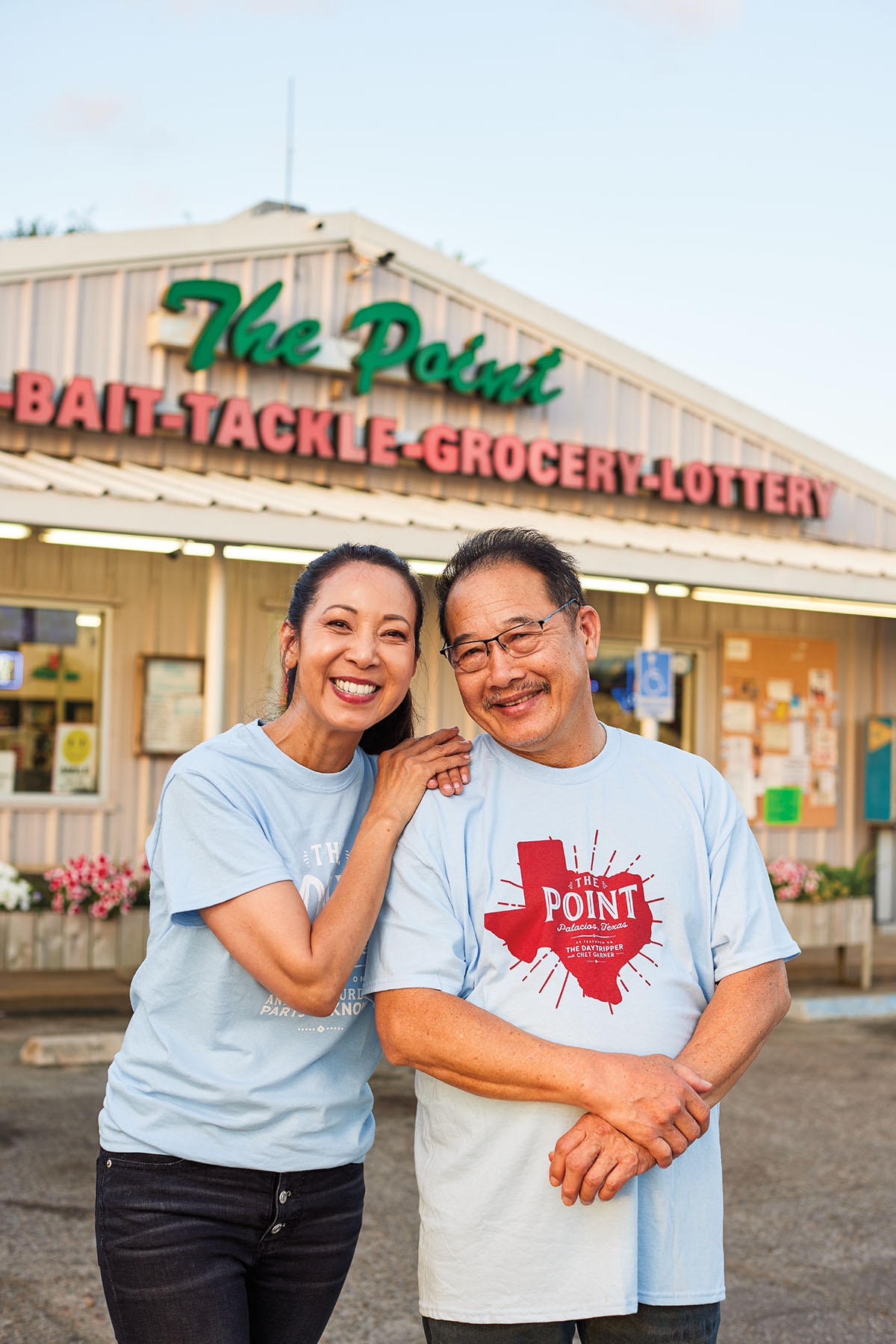 A man and woman stand in light blue t-shirts in front of a store wiwth a sign reading "The Point: Bait, Tackle, Grocery, Lottery"