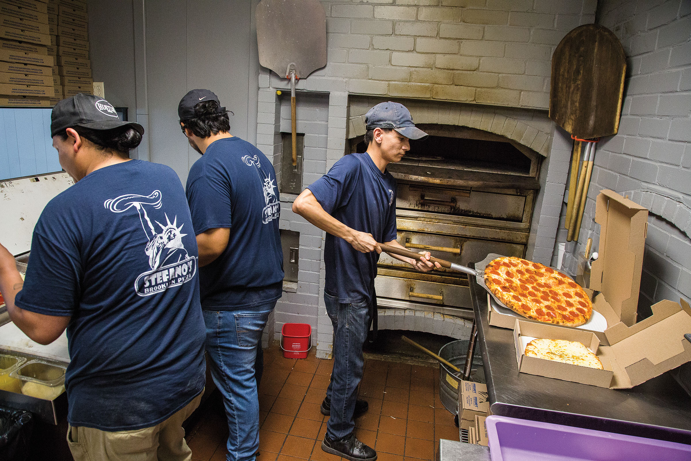 An employee takes a pizza off of a peel in front of a large oven while other employees look nearby