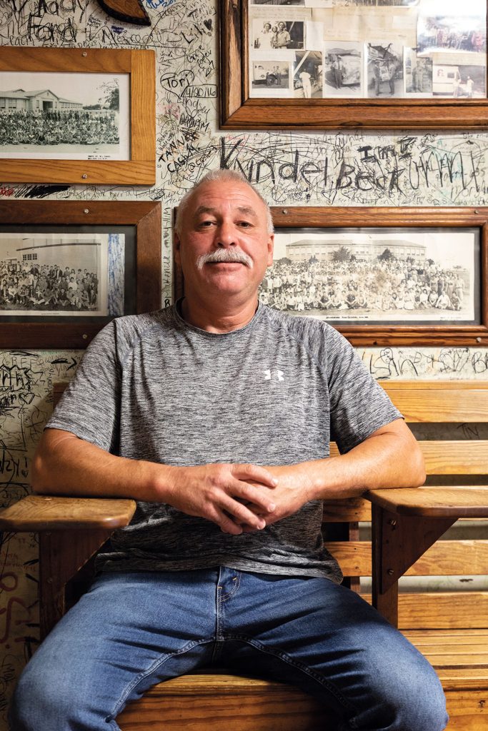 A man in a gray t-shirt and blue jeans sits in front of a wall of pictures