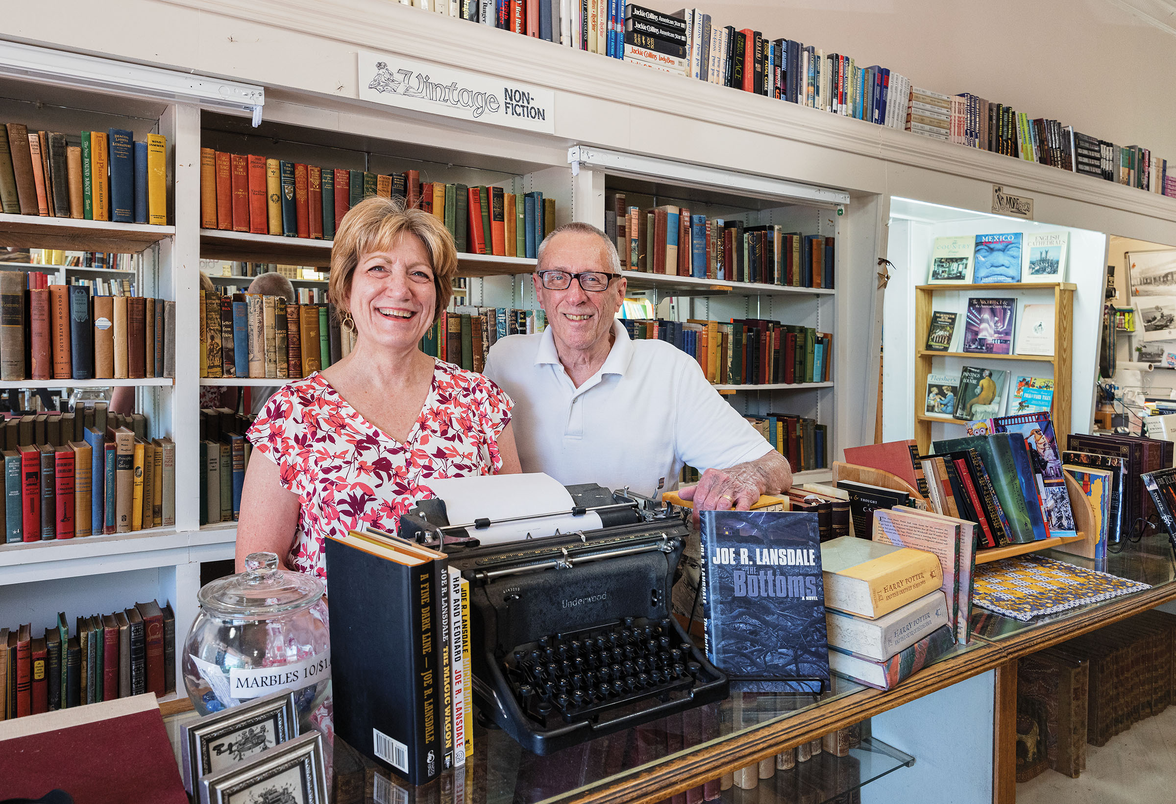 Two people smile in front of a wall of a number of colorful books