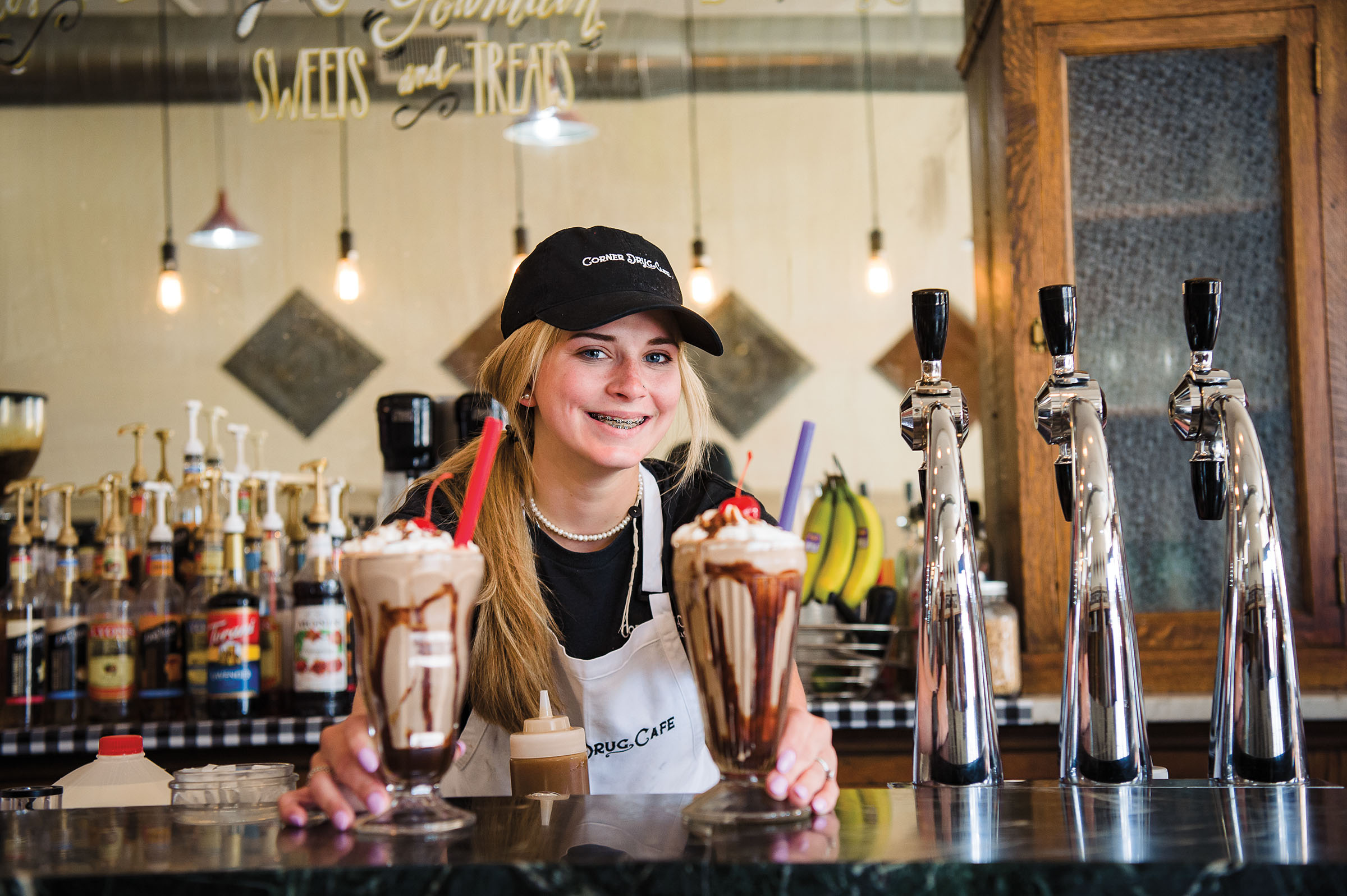 A young woman in a black baseball cap stands behind two milkshakes at a soda fountain