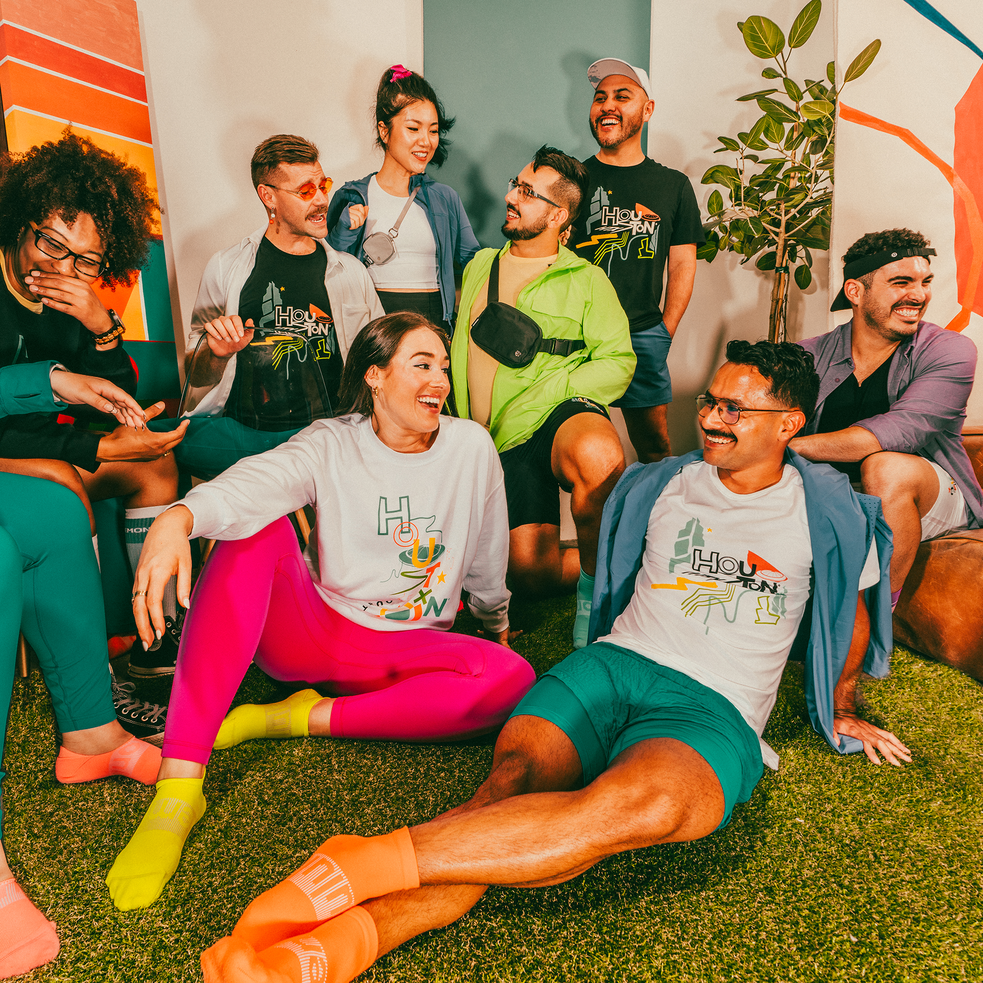 Eight people sit haphazardly on floors and couches smiling and having fun. They are all wearing shirts or shorts with the Houston design on them, and the colors are vibrant.