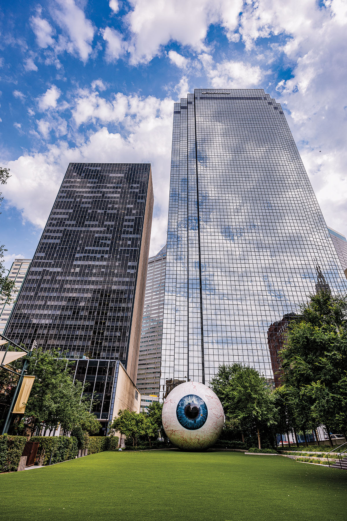 A large fiberglass eye sculpture on green grass beneath tall glass buildings