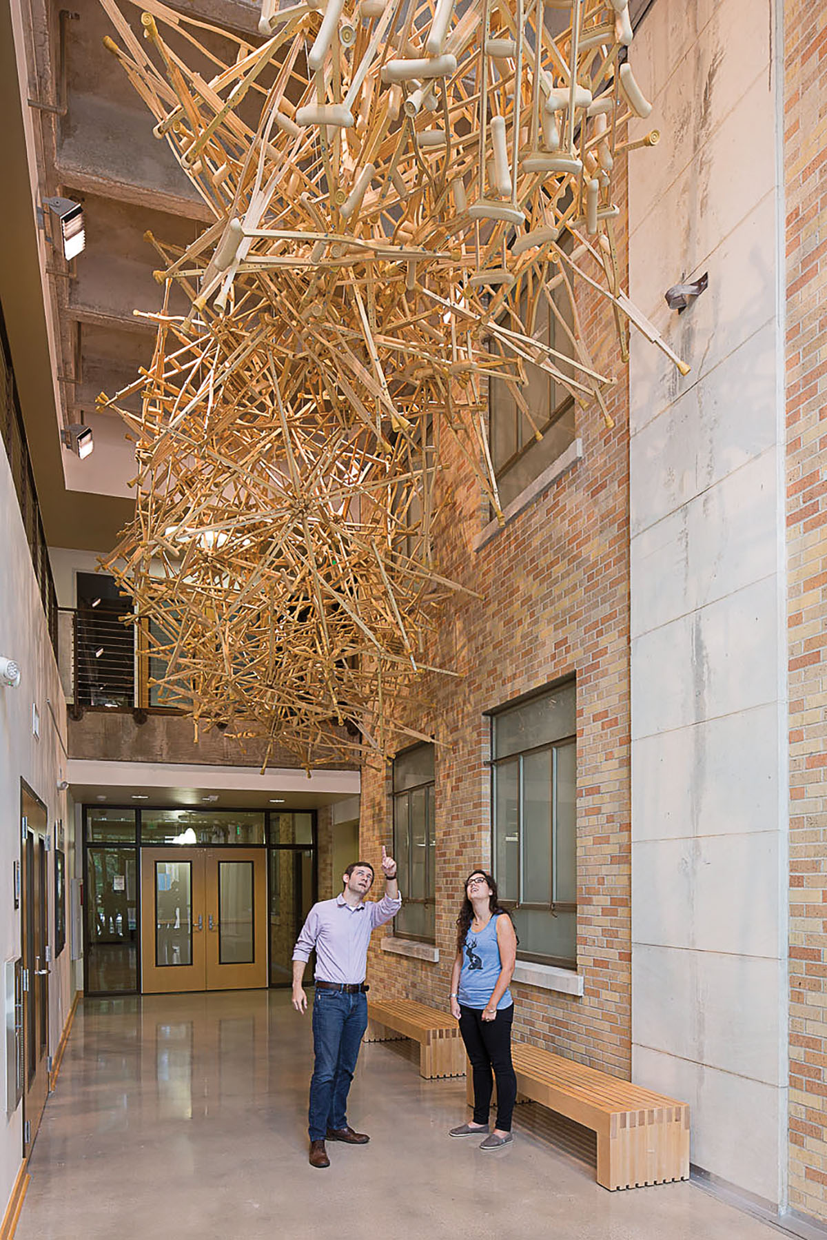 Two people stand in a hallway underneath a large and spindly sculpture made of wood