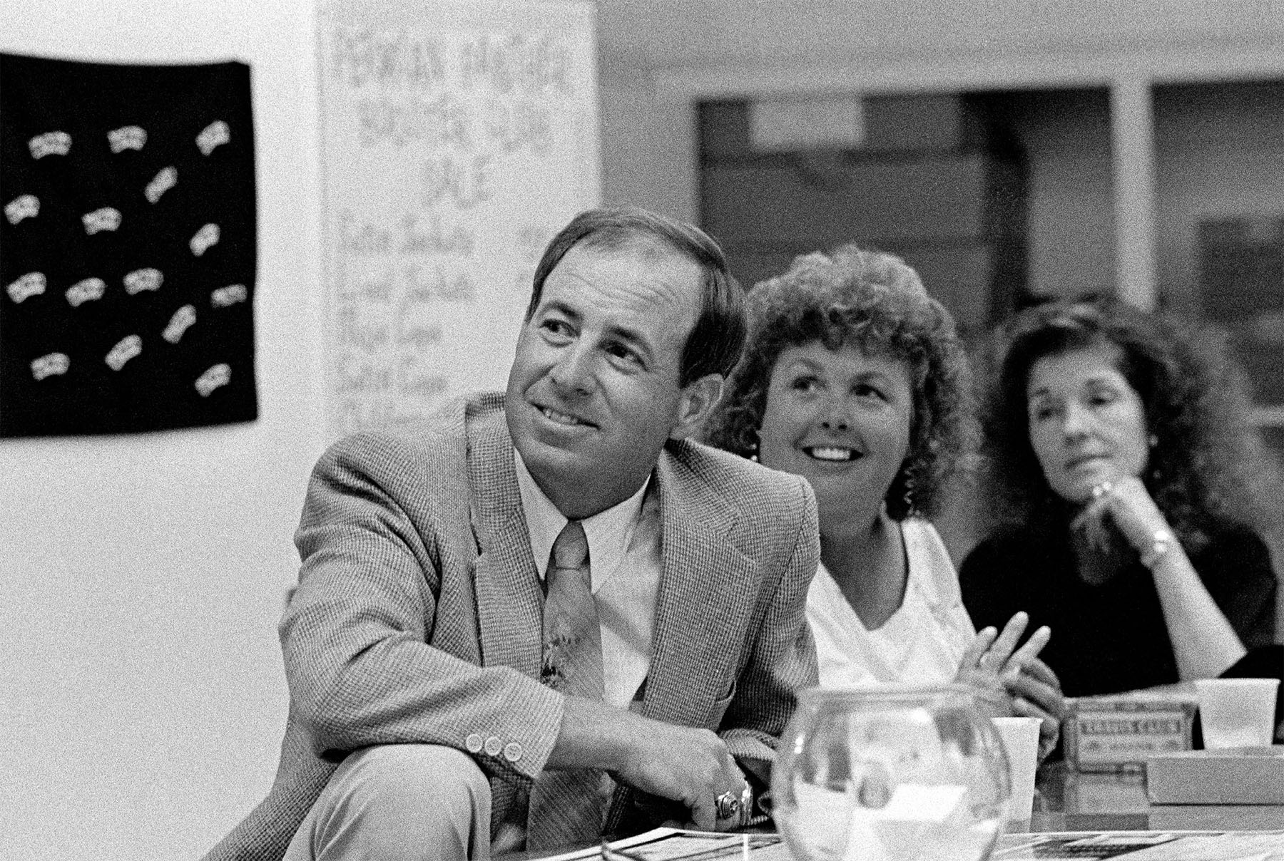 A black and white photograph of a man in a suit looking off into the distance, seated at a table next to two people 