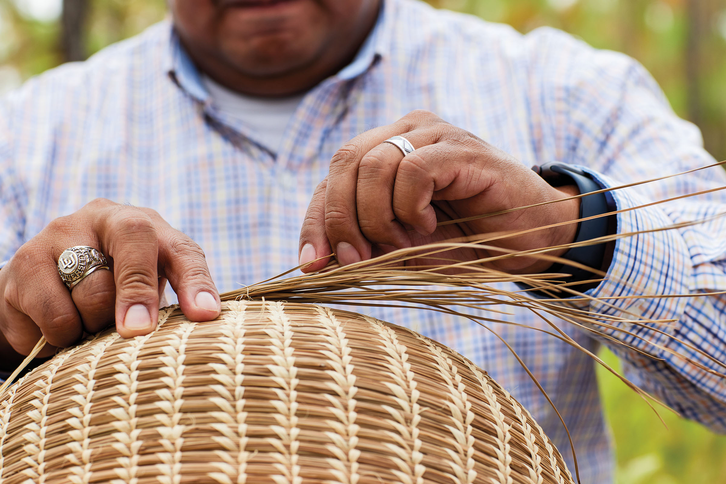 A person's hands carefully weave pieces of tan straw into the shape of a basket