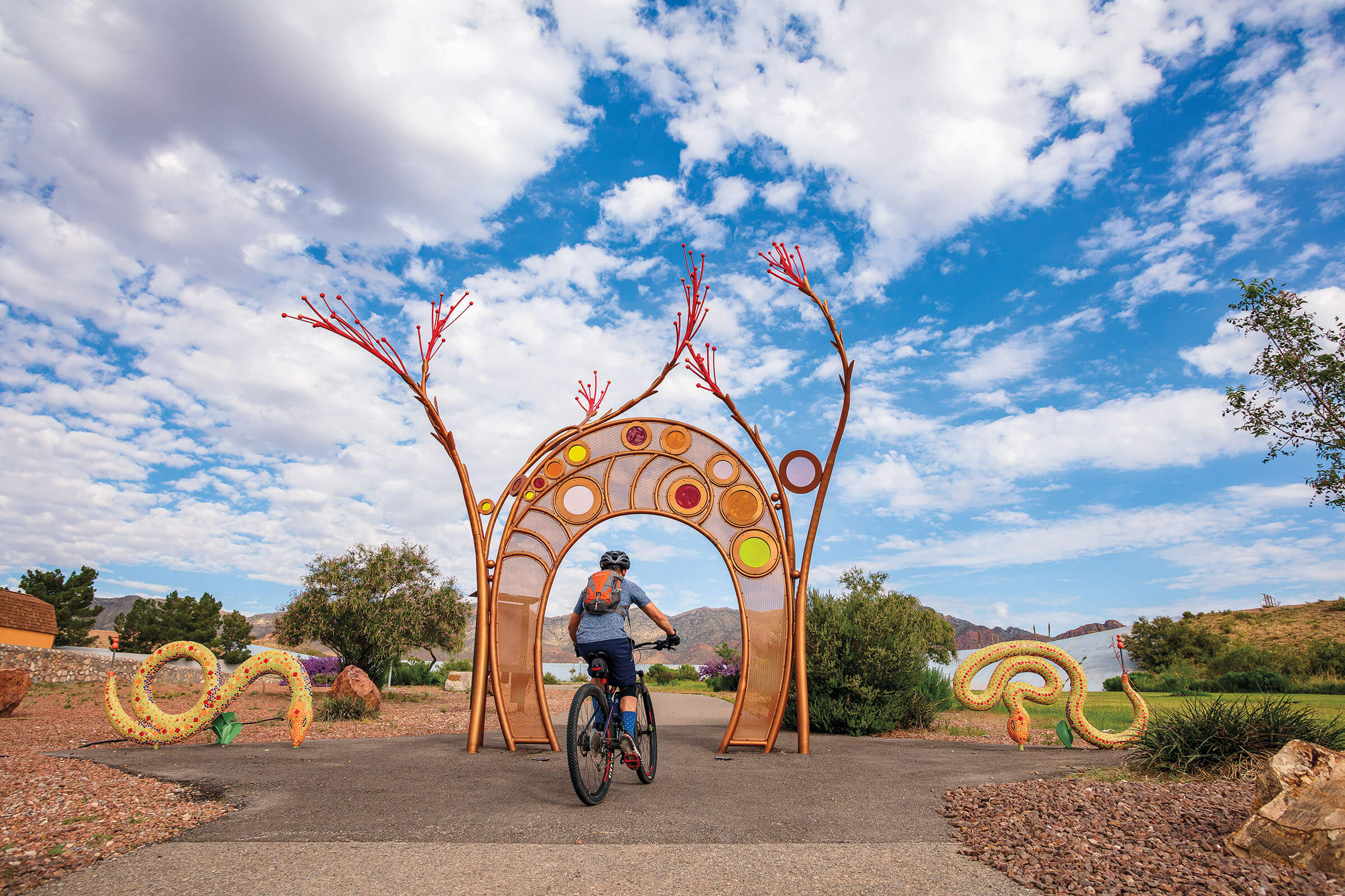 Brightly colored sculptures in red and gold making a snake and archway under blue sky. A cyclist pedals through the archway