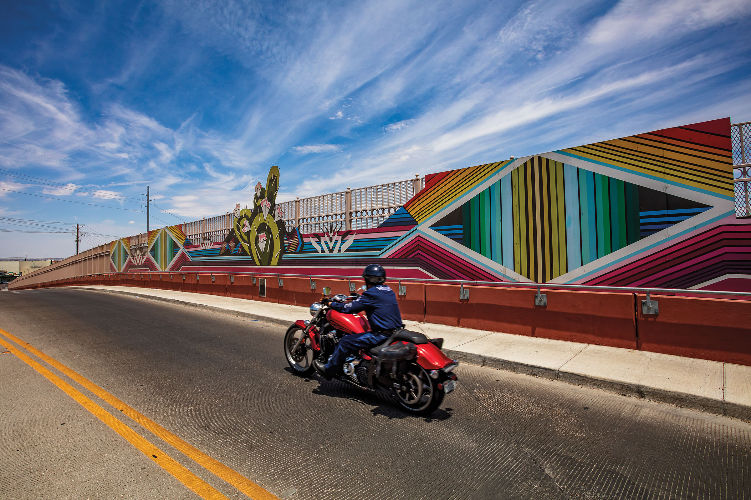 A motorcycle drives by a mural-like sculpture with bright colors along the side of a bridge