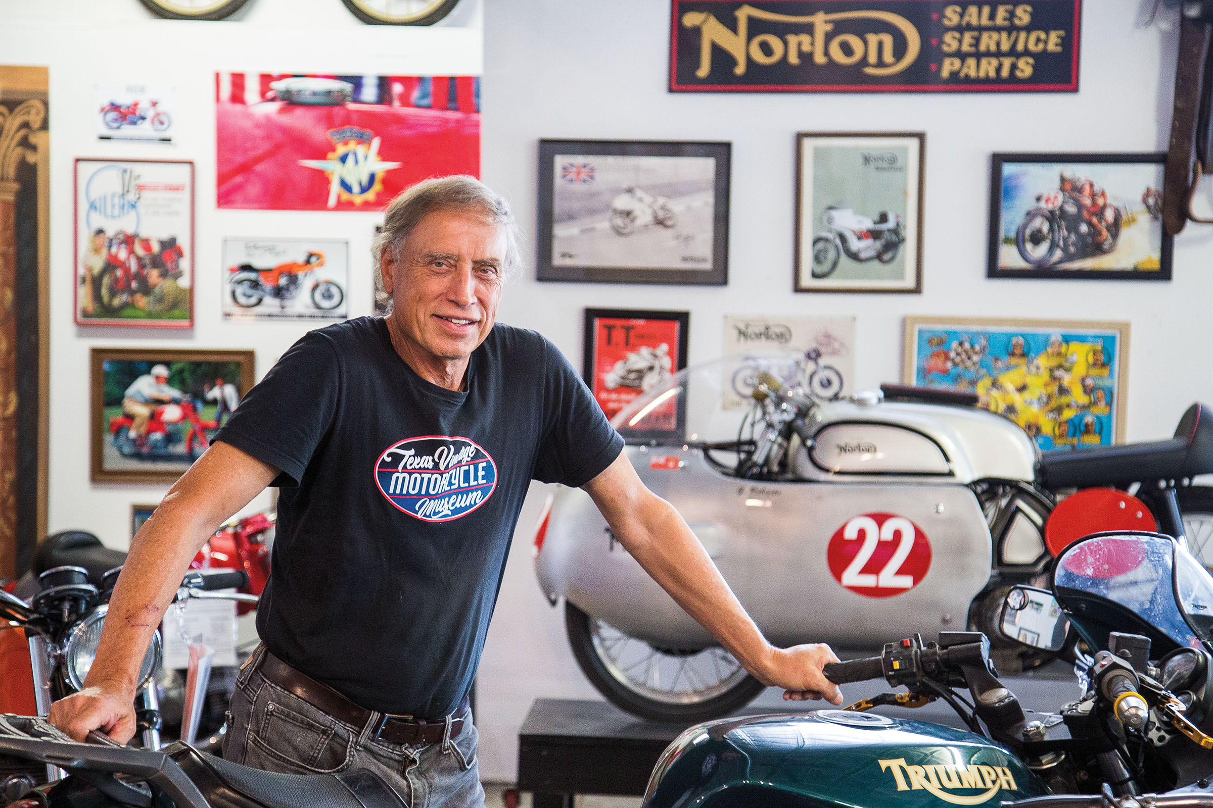 A man in a black t-shirt stands in front of numerous vintage motorcycles and memorabilia