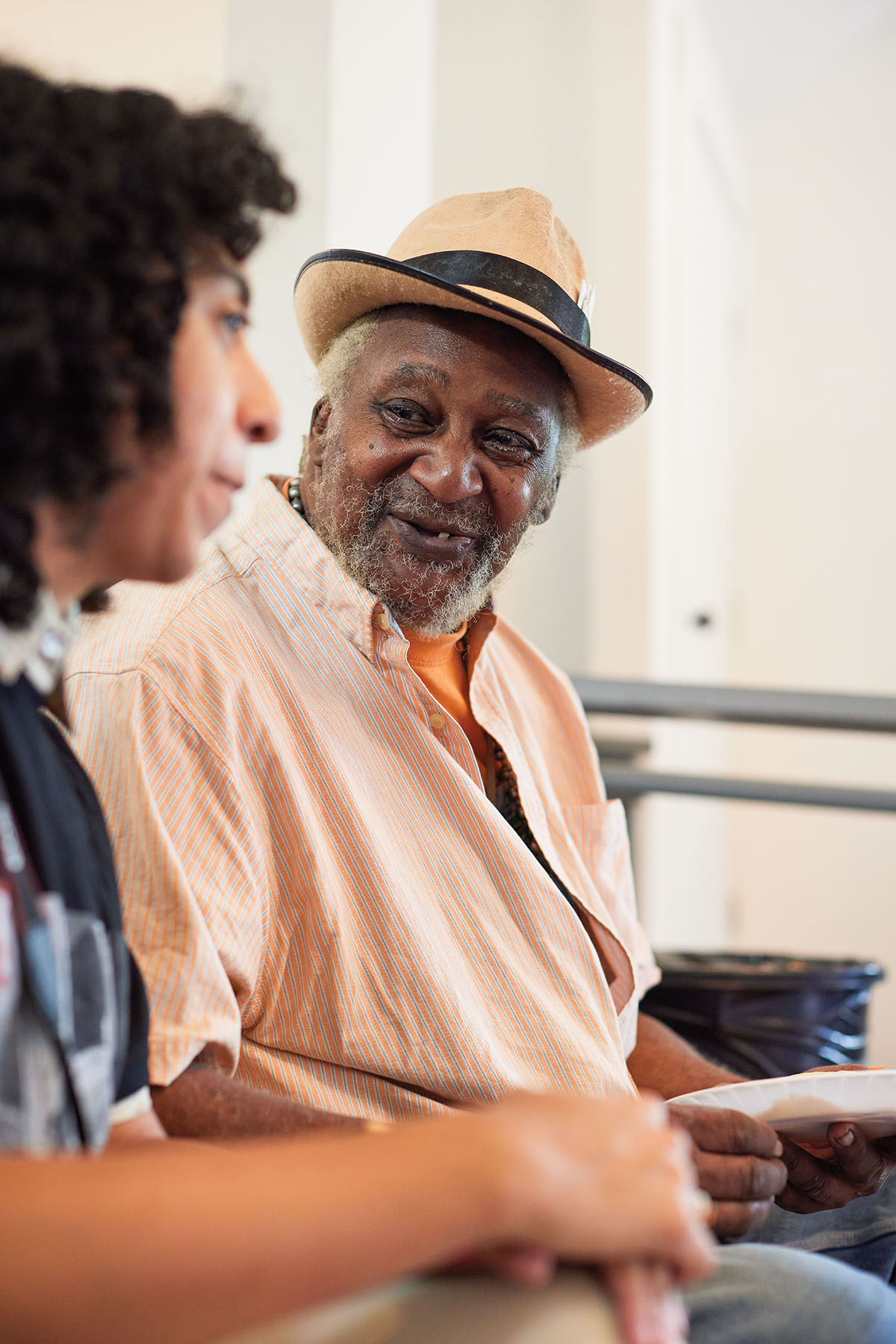 A man in a peach colored shirt and straw hat sits talking with a second person slightly blurred in the frame