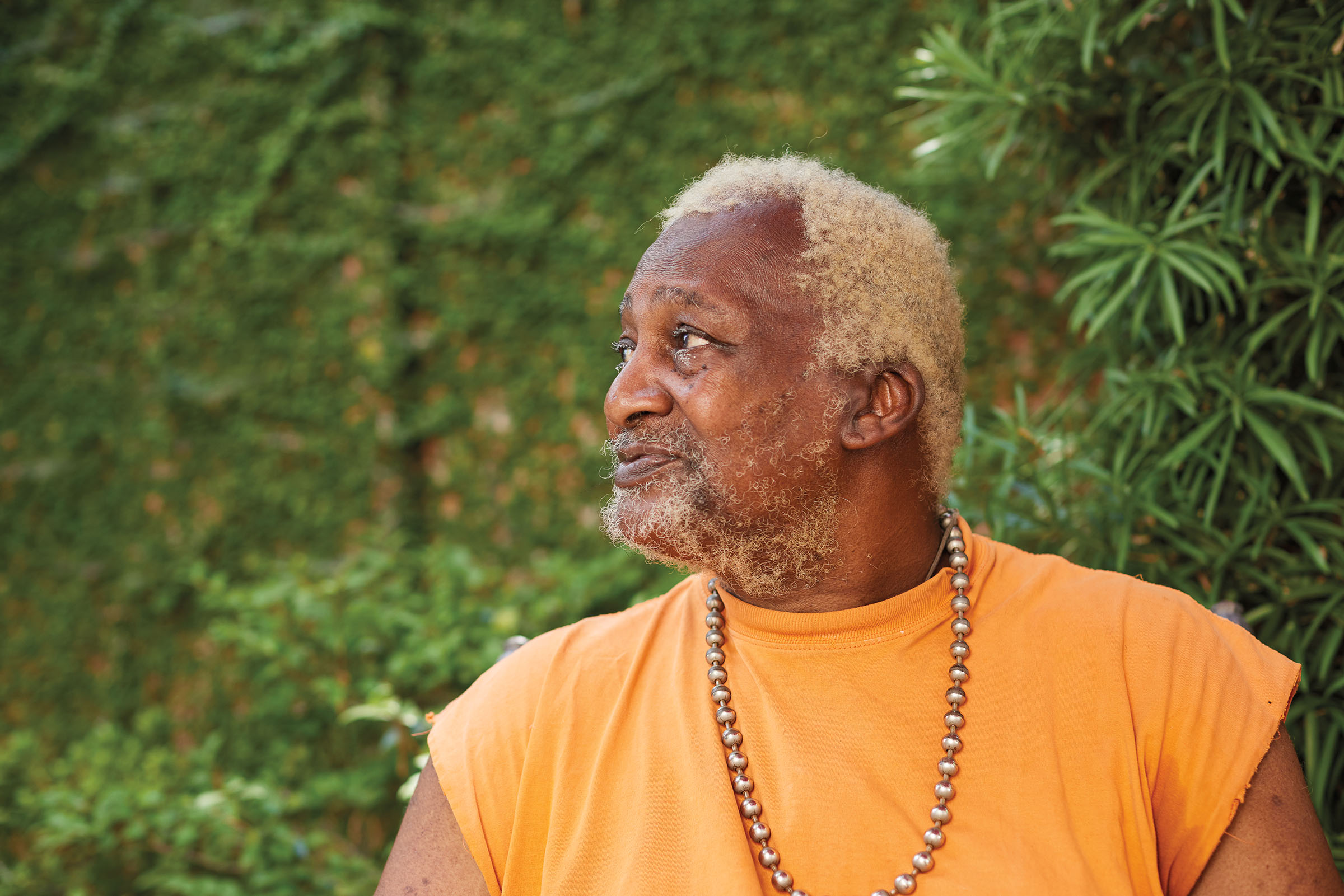 A man in an orange shirt and gold chain looks off into the distance in front of a green forested background