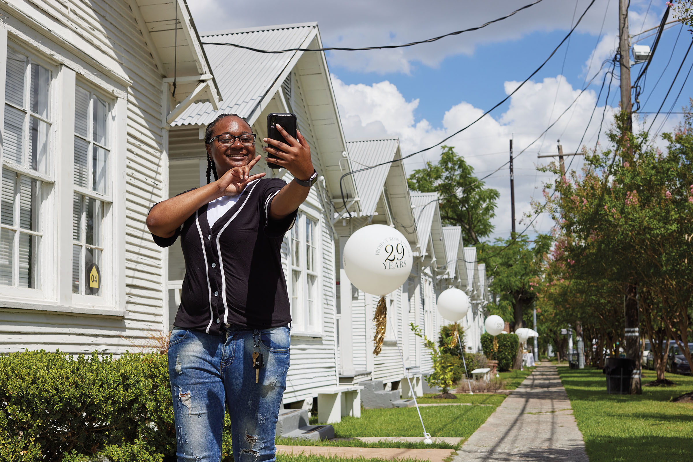 A woman in a black t-shirt and jeans takes a selfie in front of a collection of uniform white houses under blue sky