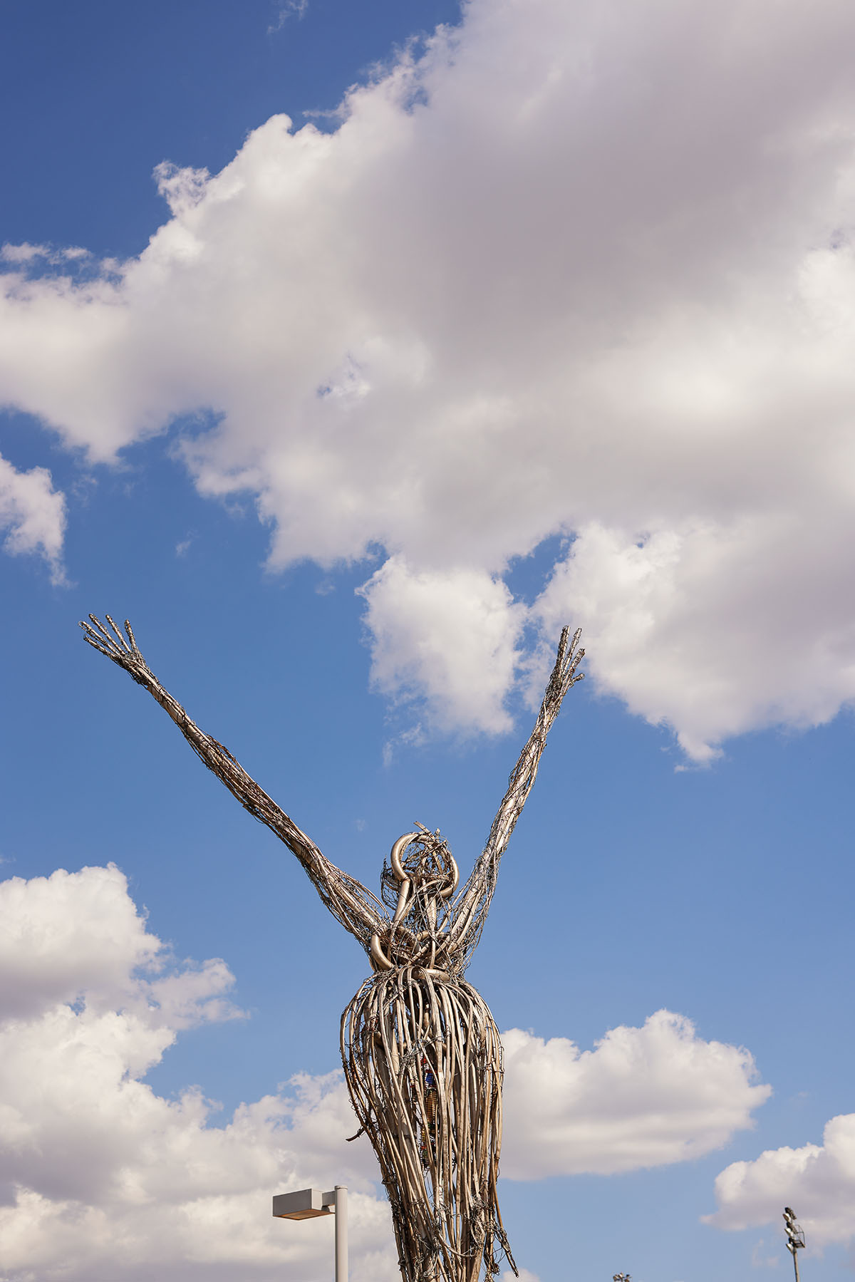 A tall sculpture made of pieces of metal shaped to look like a figure reaching up its arms in front of a blue sky