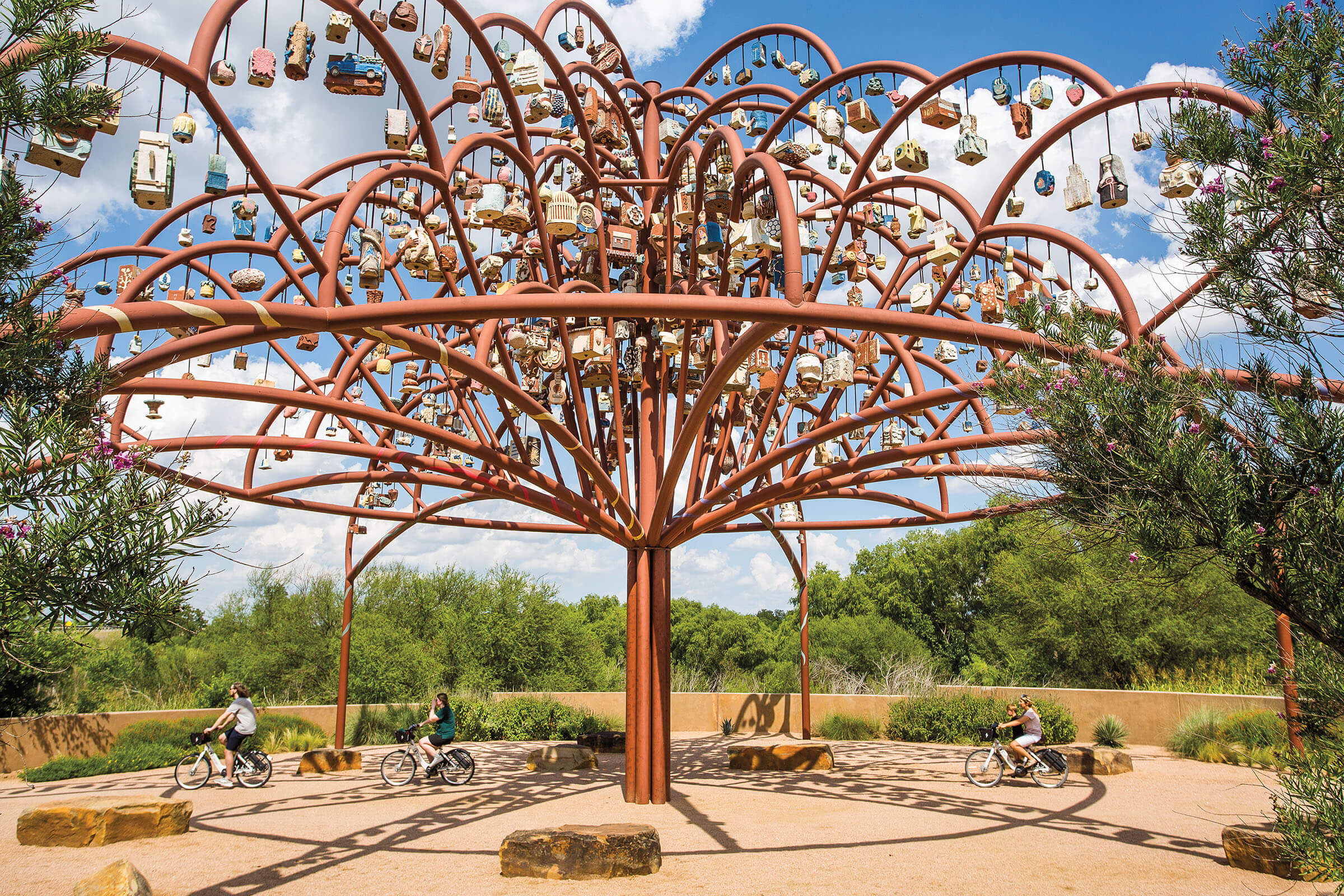 A rust-red tree sculpture filled with decorative ornaments on dusty ground under blue sky