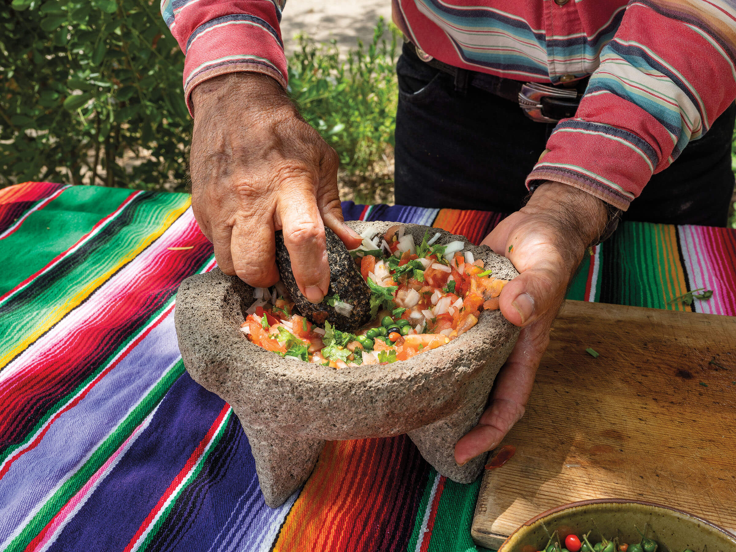 A man's hands mashes tomato, chile, and onion in a molcajete to make salsa