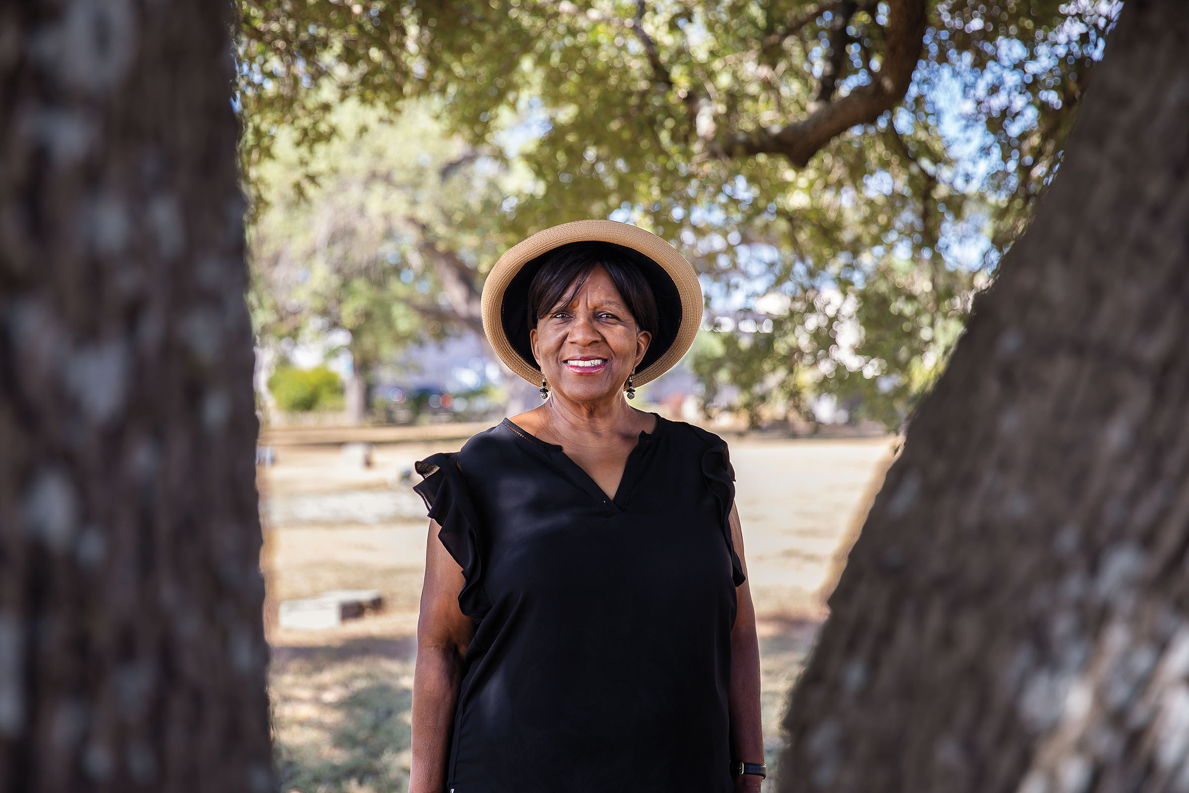 A woman in a black shirt and straw hat stands in front of two green trees in a cemetery
