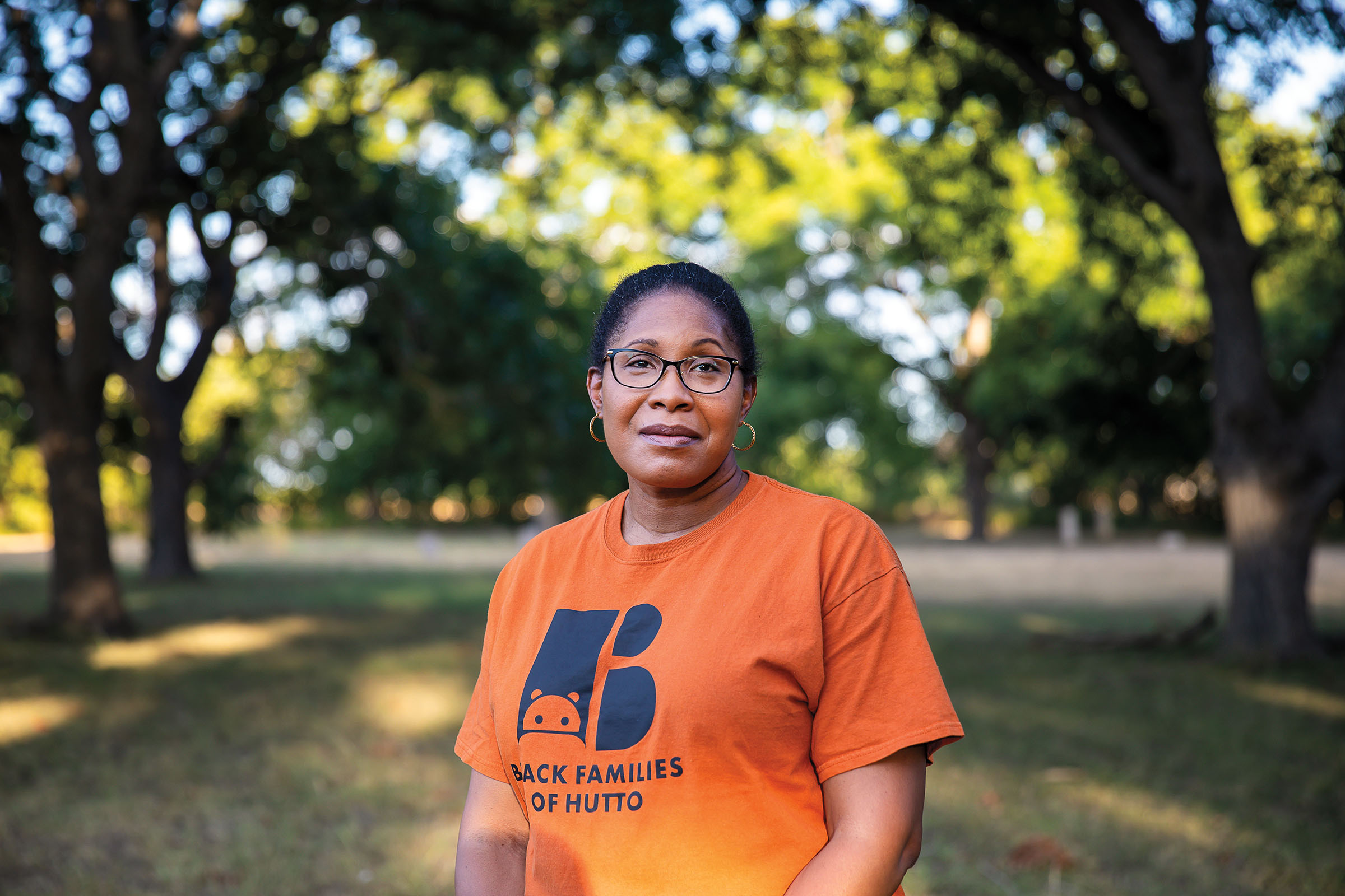 A woman in an orange shirt stands in front of a canopy of green trees