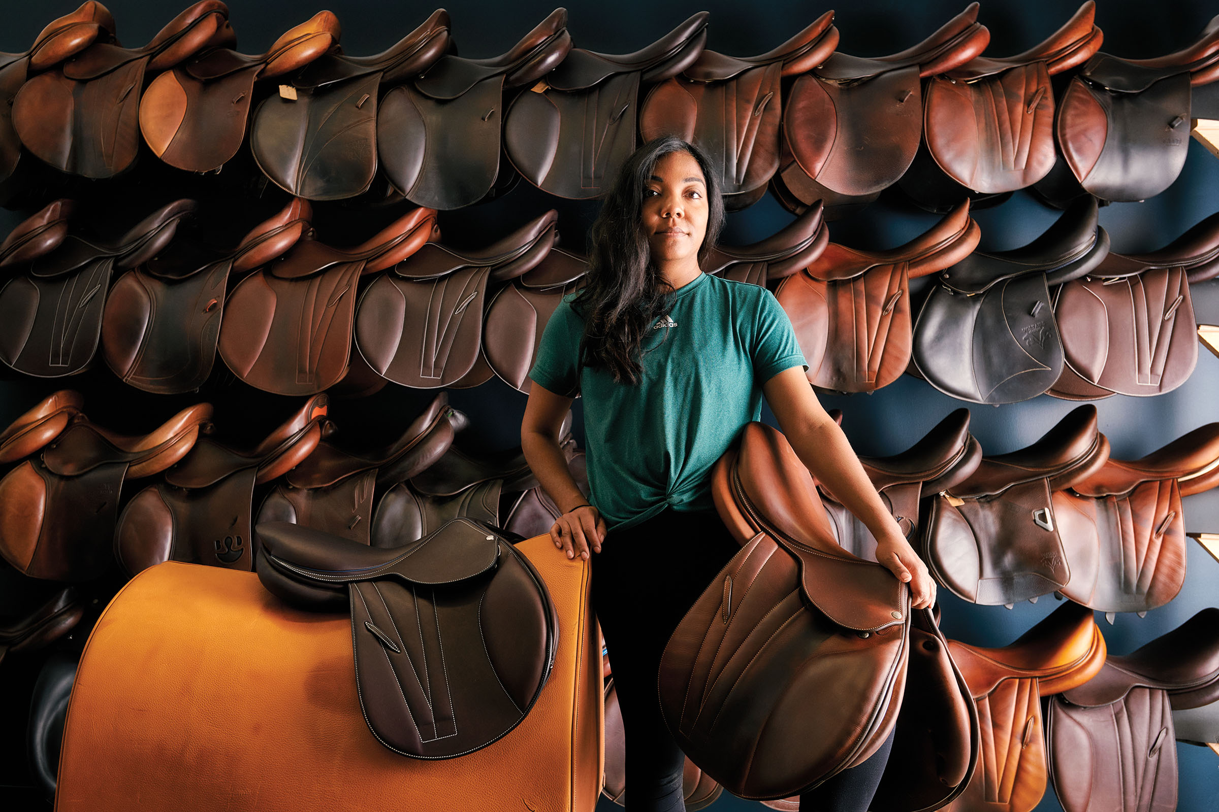 A woman stands with a dark leather saddle in front of a wall with numerous rich leather saddles in various shades of brown