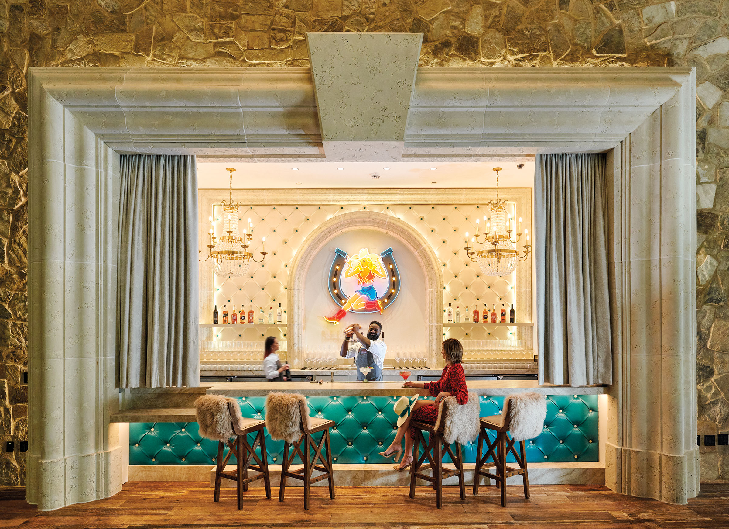 A bartender shakes a drink in front of a well-lit bar with bright turquoise barstools and a customer sitting