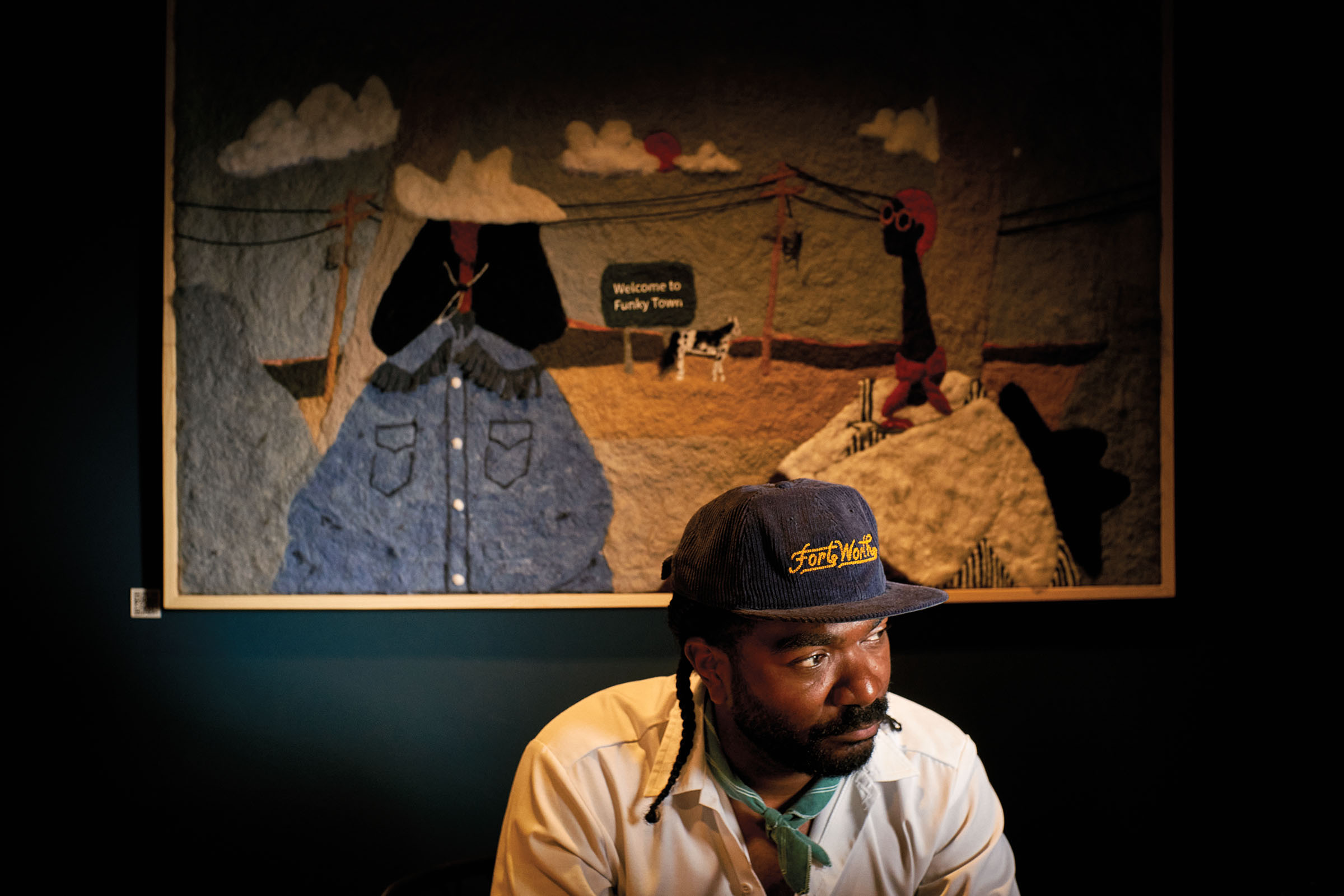 A man in a ball cap and white shirt sits in front of a mural in a dimly-lit room