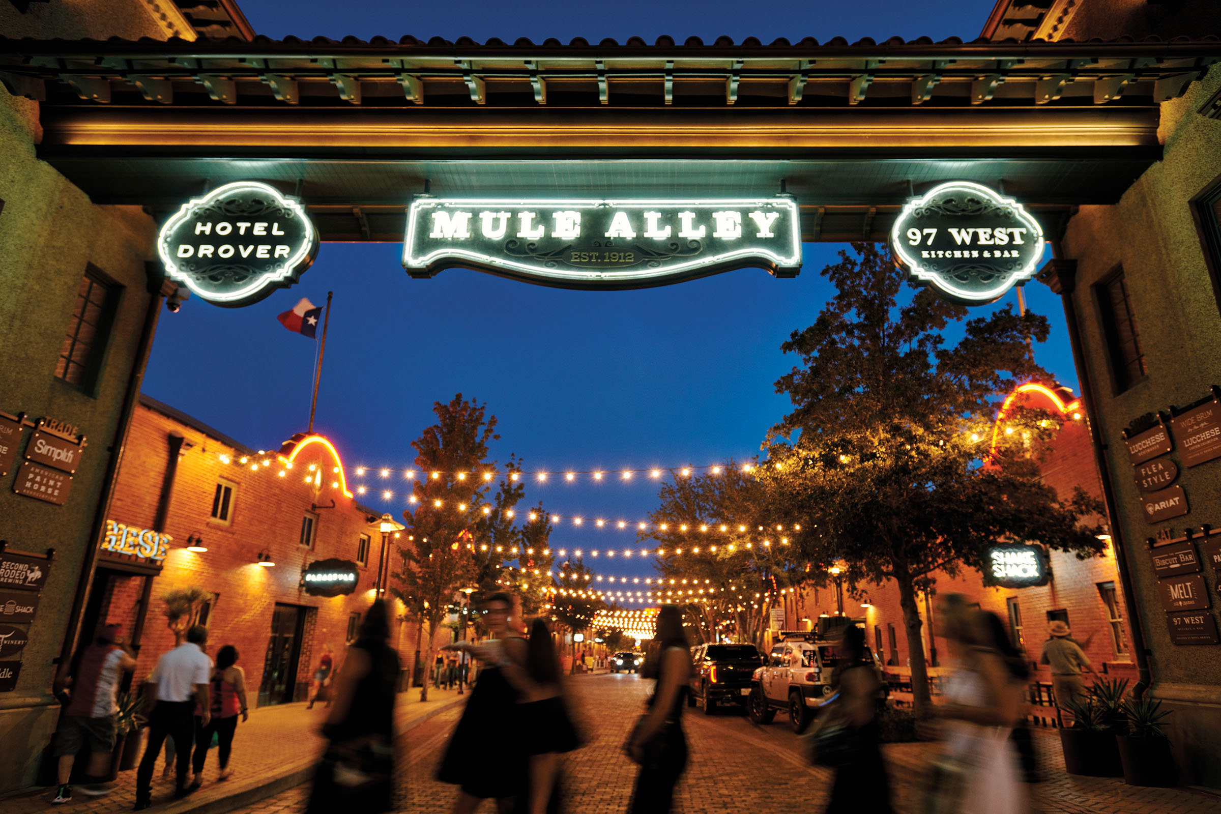 Bright string lights and neon signs illuminate a corridor filled with people milling about in an evening scene