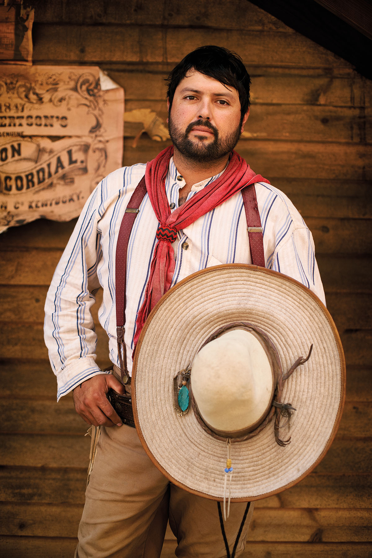 A vaquero stands holding his hat in front of a wood-paneled wall