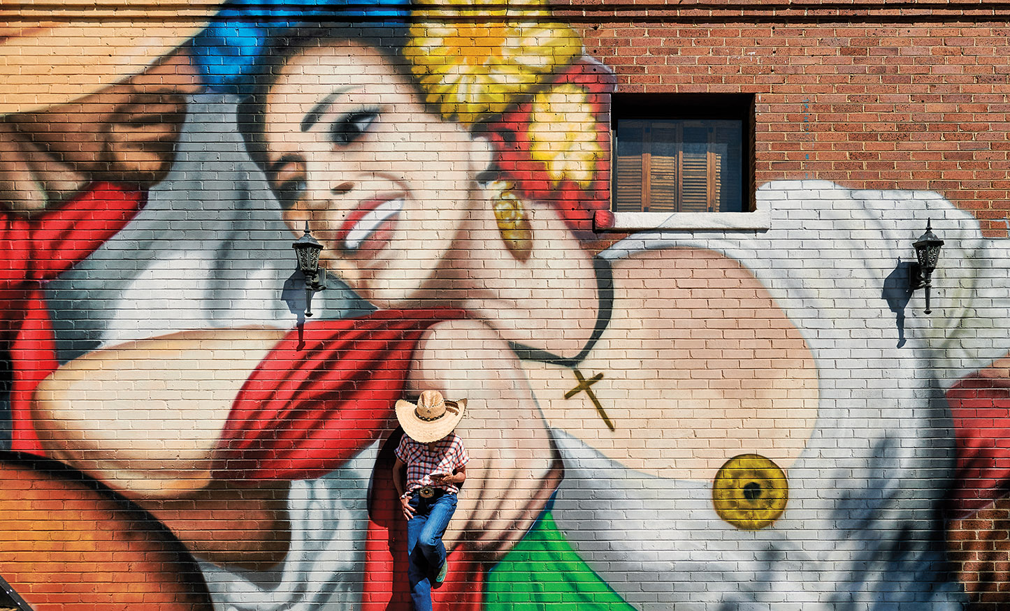 A person in a cowboy hat leans with their head down and back against a large colorful mural depicting a dancing woman
