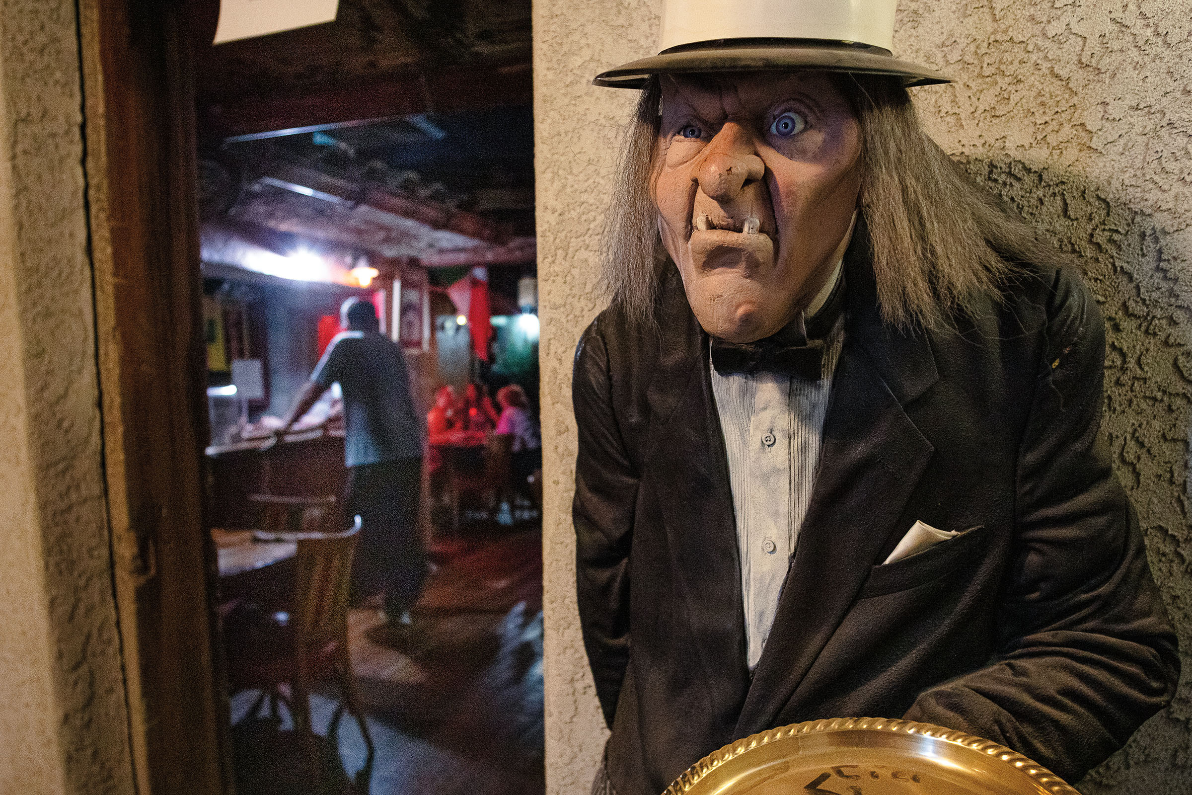 A wax sculpture of a scary-looking man with long hair and a hat near the exterior of a room