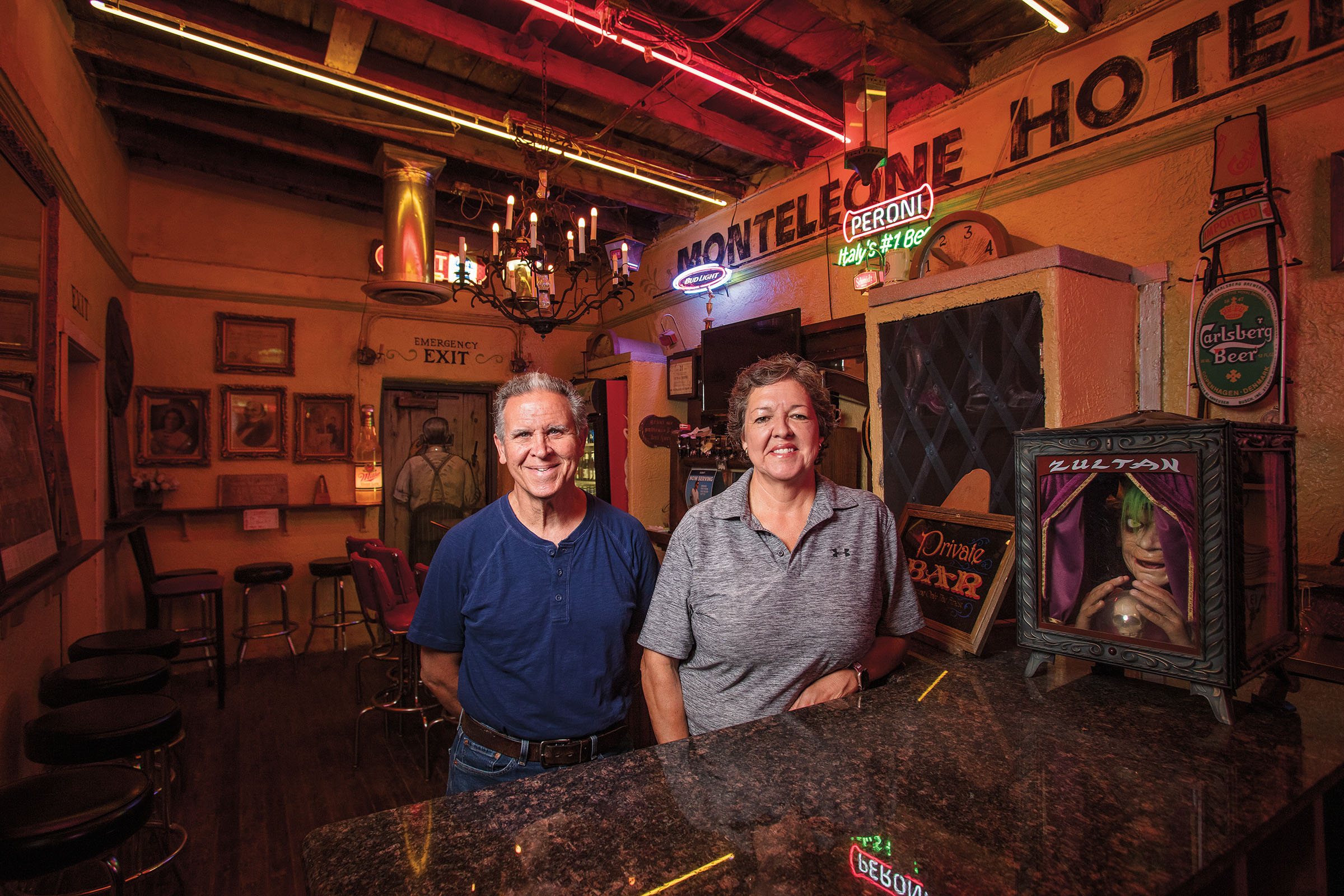 A man and woman stand behind a granite bar inside of an old-fashioned bulding with a candle chandelier and neon signs