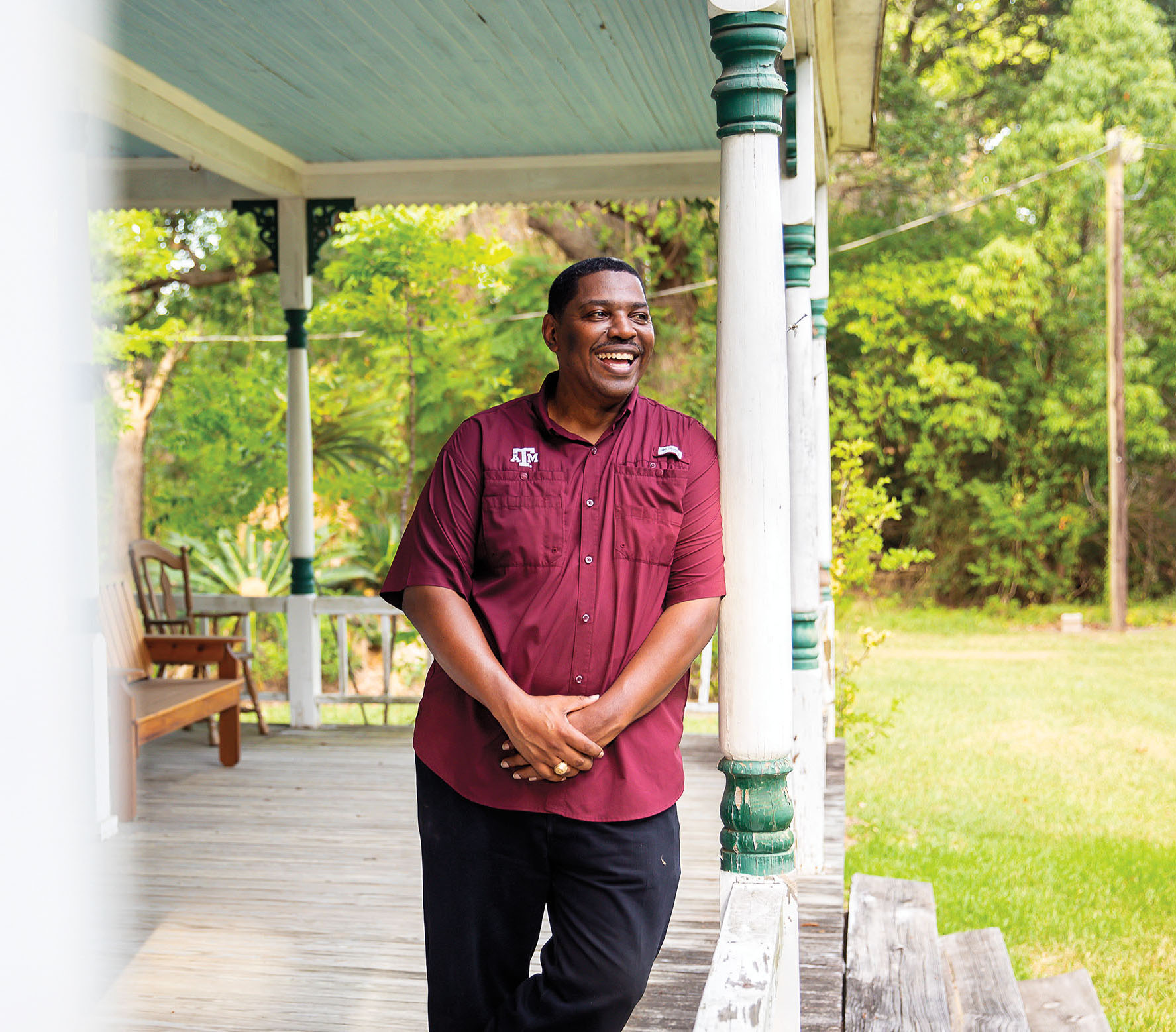 A man in a maroon Texas A&M shirt leans against a white post on a porch in a lush green area