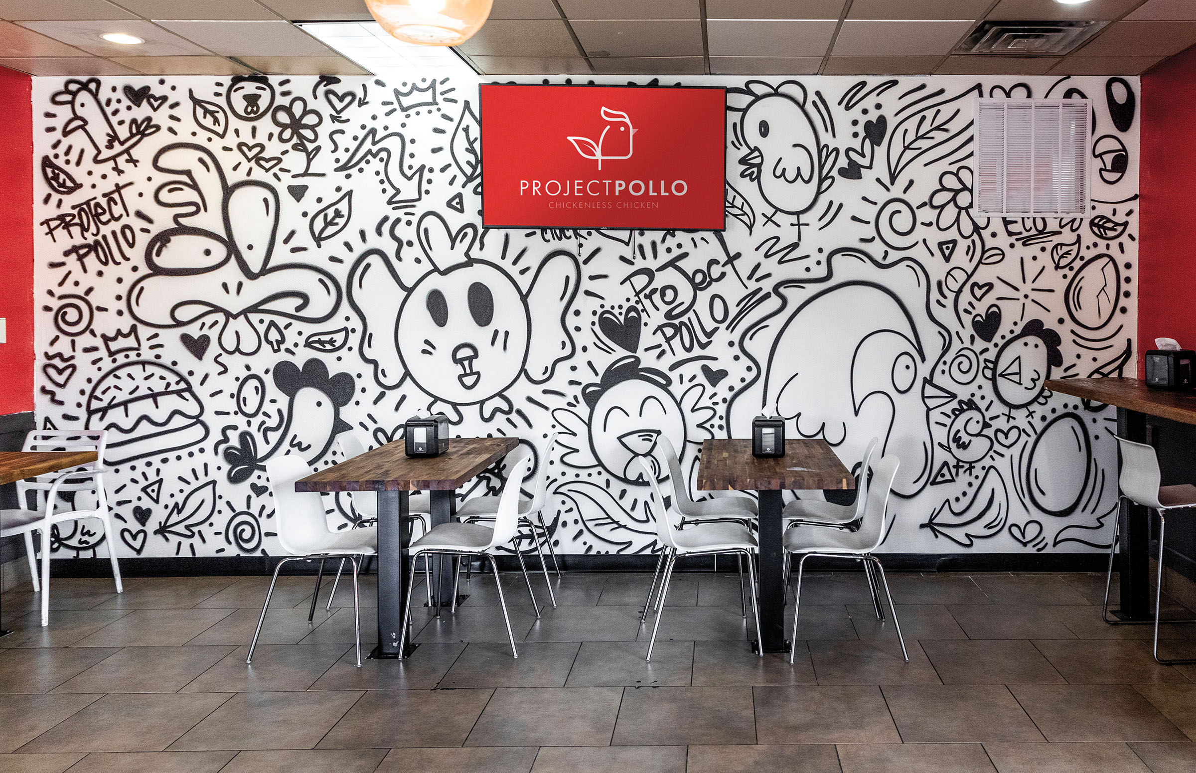 A zany mural in black and white adorns the wall of a building behind a bright red sign reading Project Pollo. Wooden tables and metal chairs dot the foreground.
