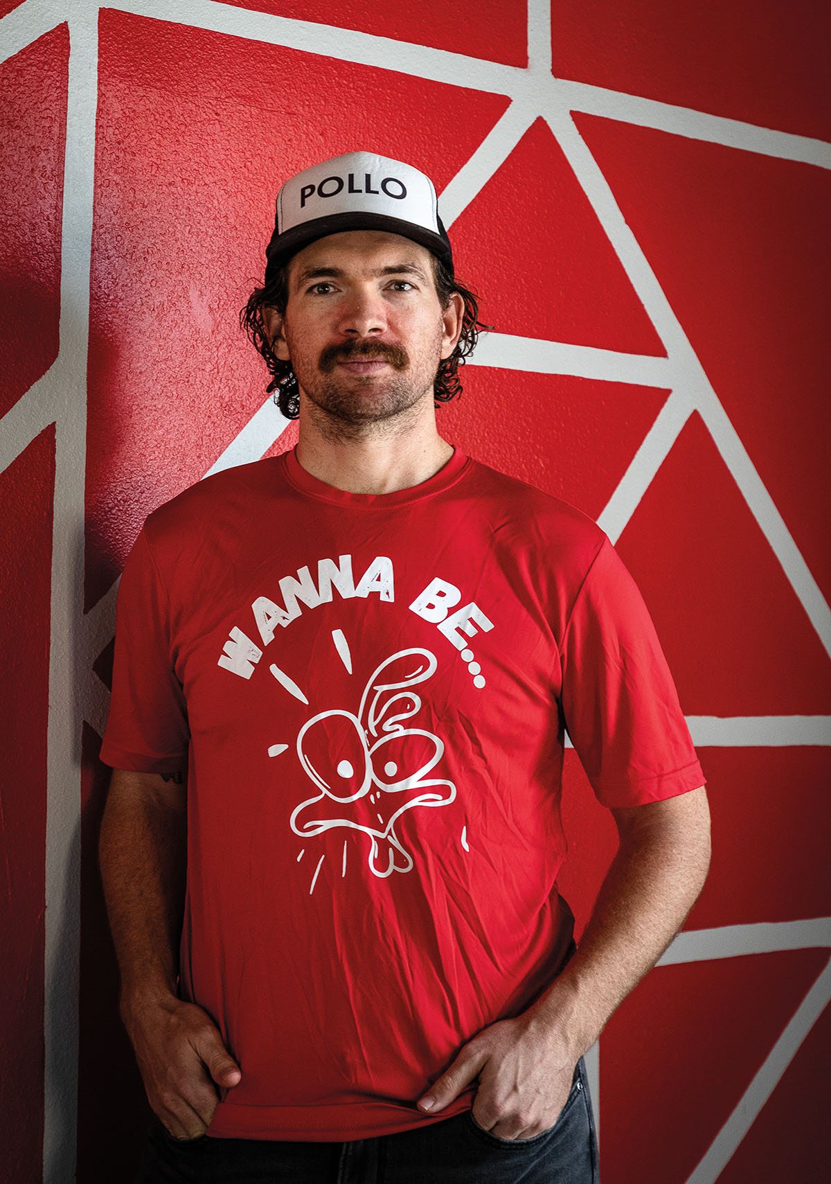 A man in a bright red shirt reading "Wanna Be" stands with a somber expression in front of a bright red wall wearing a white baseball cap