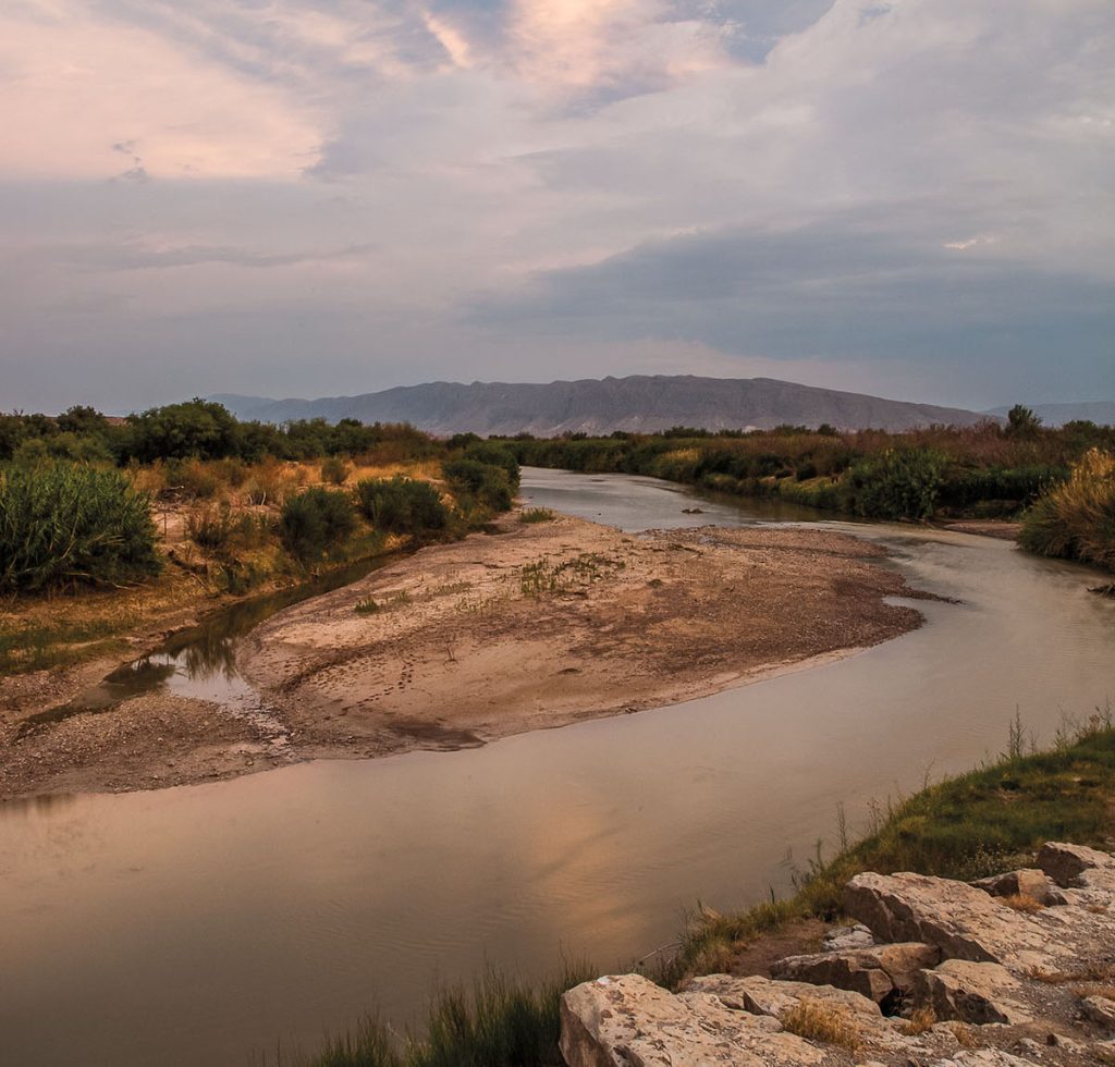 The gray-blue waters of the Rio Grande cut through a swath of desert landscape with a tall mountain in the background