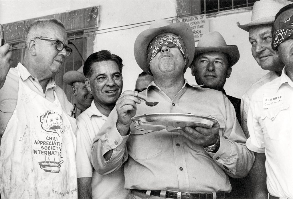 A group of men stand around a blindfolded man holding a plate of chili and a spoon