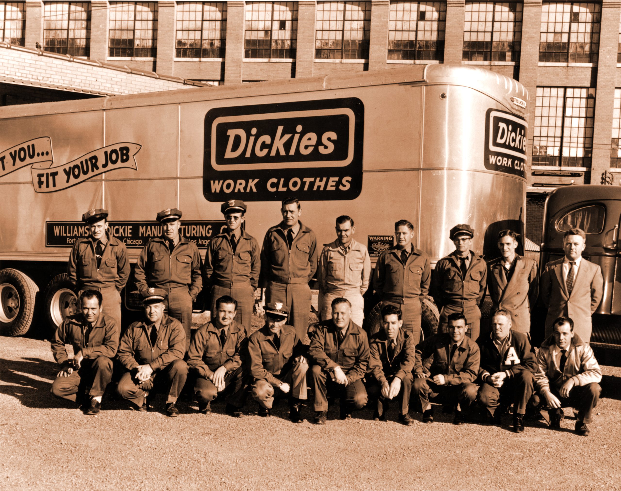 The black-and-white photo from the 1940s shows truckers posing in front of a Dickies truck. The truckers form two lines, one with men standing in the back line and then a row of men crouching down in front of them. 
