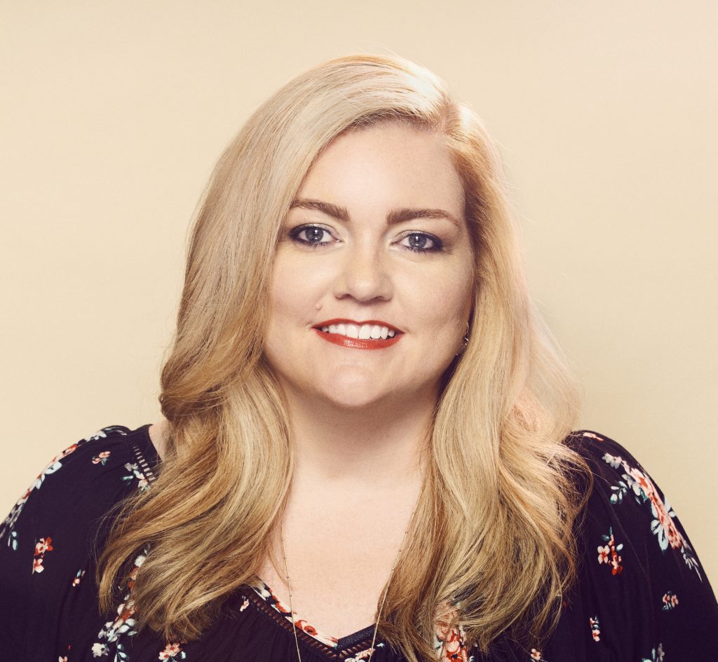 An image of a woman with blonde hair wearing a dark colored blouse and flower pattern