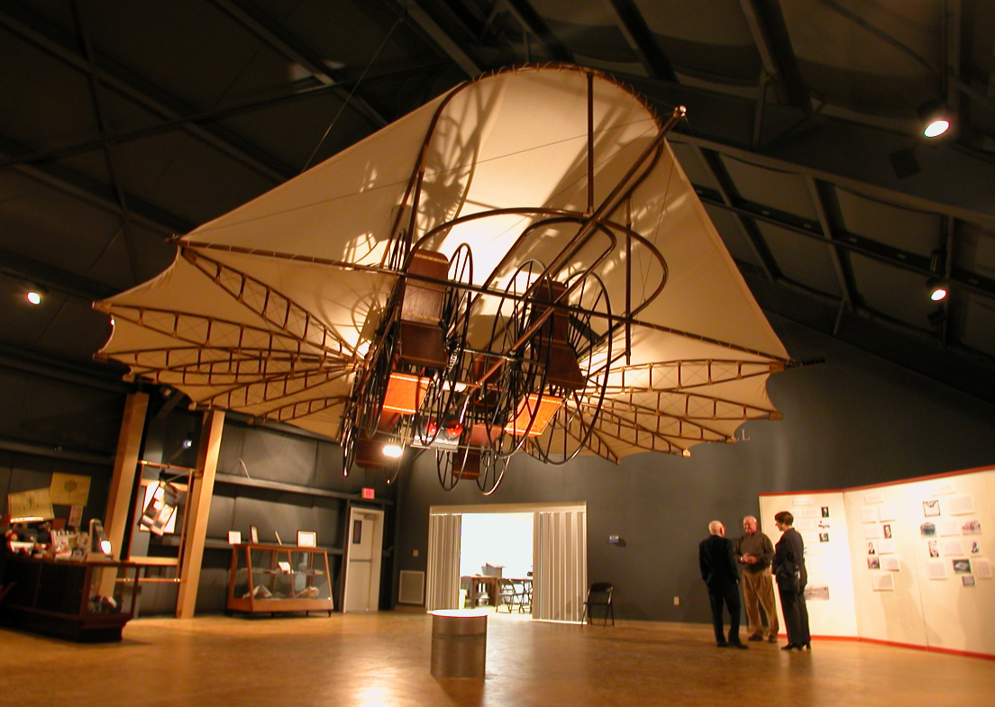 A shot of the "Ezekiel" flying machine hangs from the ceiling in an exhibition room at the Northeast Texas Rural Heritage Center and Museum in Pittsburg. Three people stand underneath the contraption, a structure that looks like a nun's cornette with wheels. 