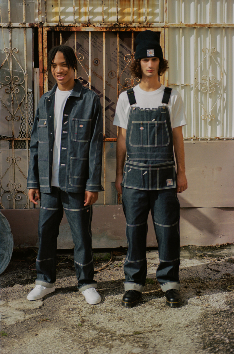 In this photo, two young men pose in Dickies denim styles. The model on the left wears jeans, a white T-shirt, and a denim jacket, while the model on the right wears denim overalls with a white T-shirt.